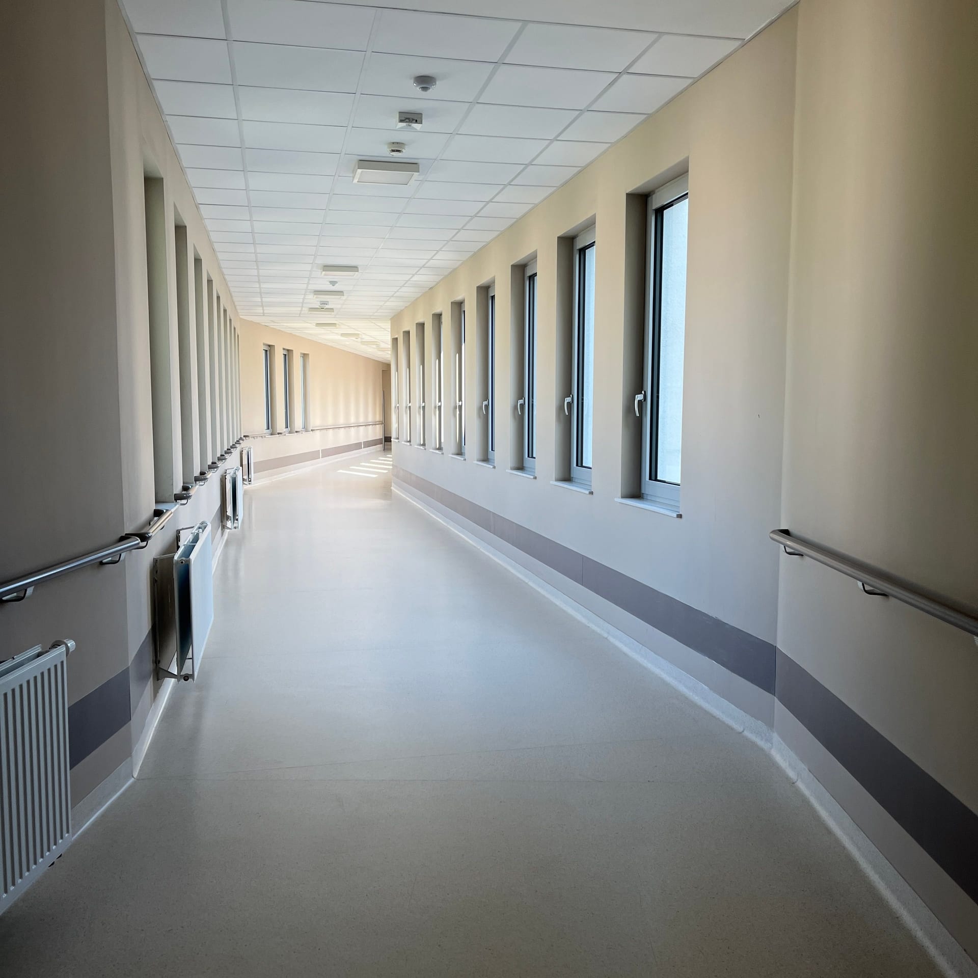 Pasillo vacío de un hospital con ventanas a un lado, pasamanos y luz natural sobre el piso claro.