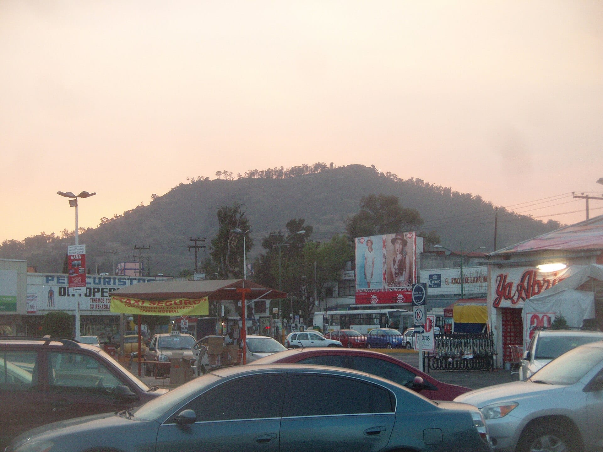 Automóviles y comercios sobre una avenida de Iztapalapa, con el Cerro de la Estrella al fondo durante el atardecer.