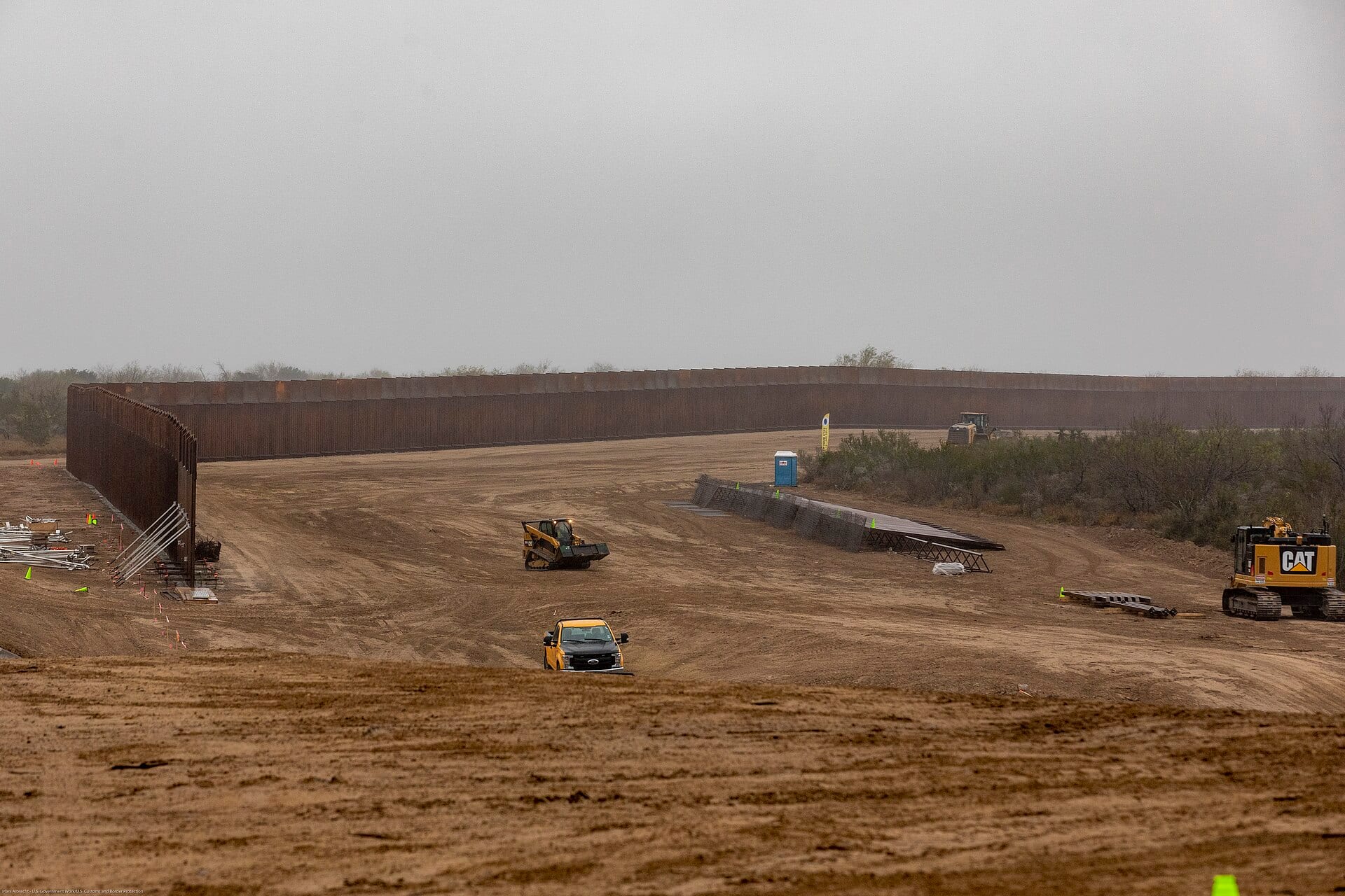 Un muro metálico alto cruza un terreno de tierra con maquinaria amarilla de construcción y una camioneta, bajo un cielo gris.