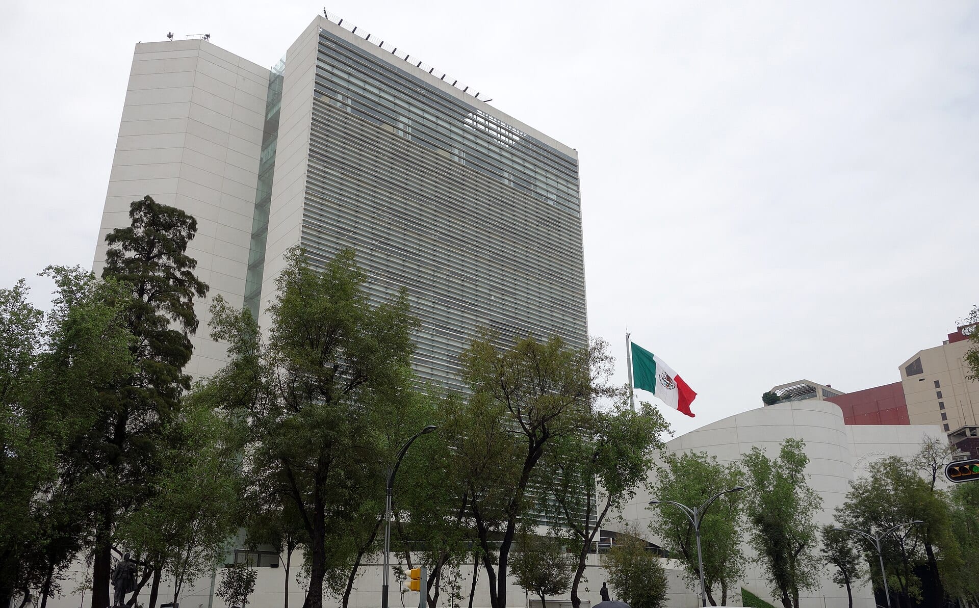 Torre blanca de fachada con celosías del Senado, rodeada de árboles, con una bandera de México ondeando a la derecha bajo un cielo nublado.