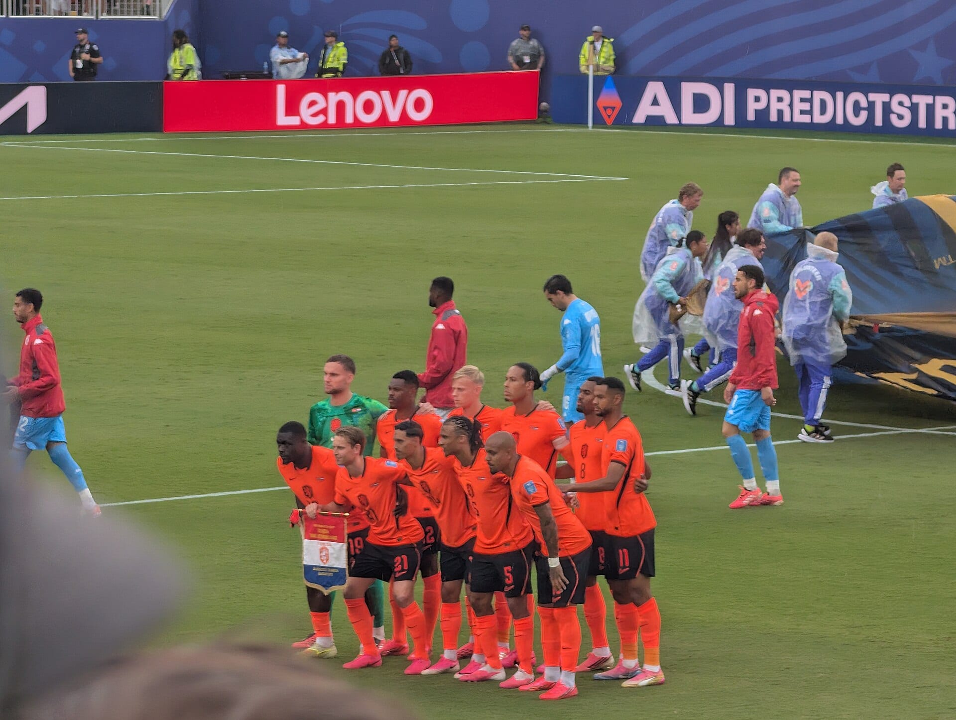Jugadores de Países Bajos con uniforme naranja posan agachados en el césped antes de un partido, mientras personal con impermeables recoge una lona al fondo.