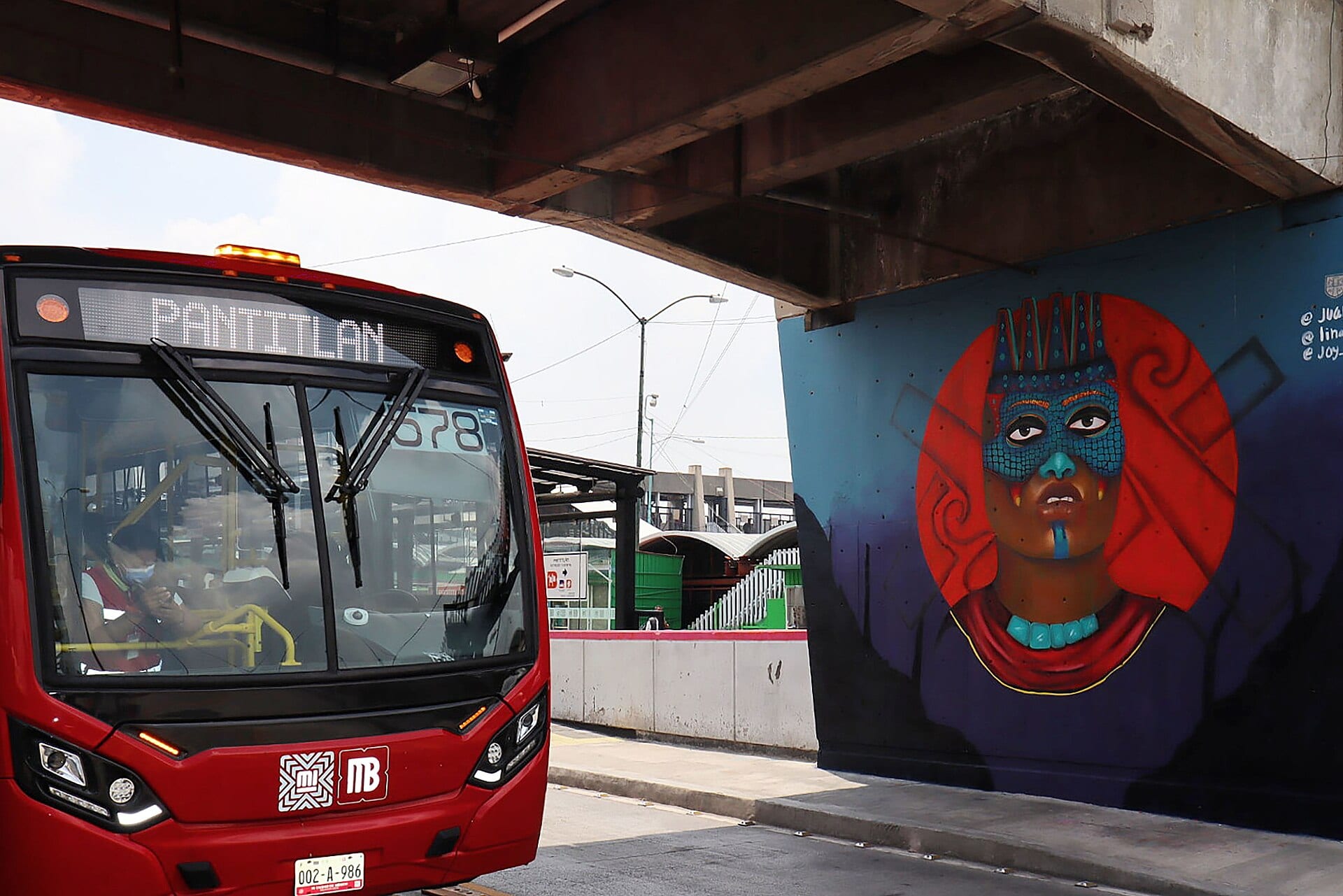 Un autobús rojo del Metrobús con destino Pantitlán junto a un mural bajo un paso elevado, con andenes al fondo.