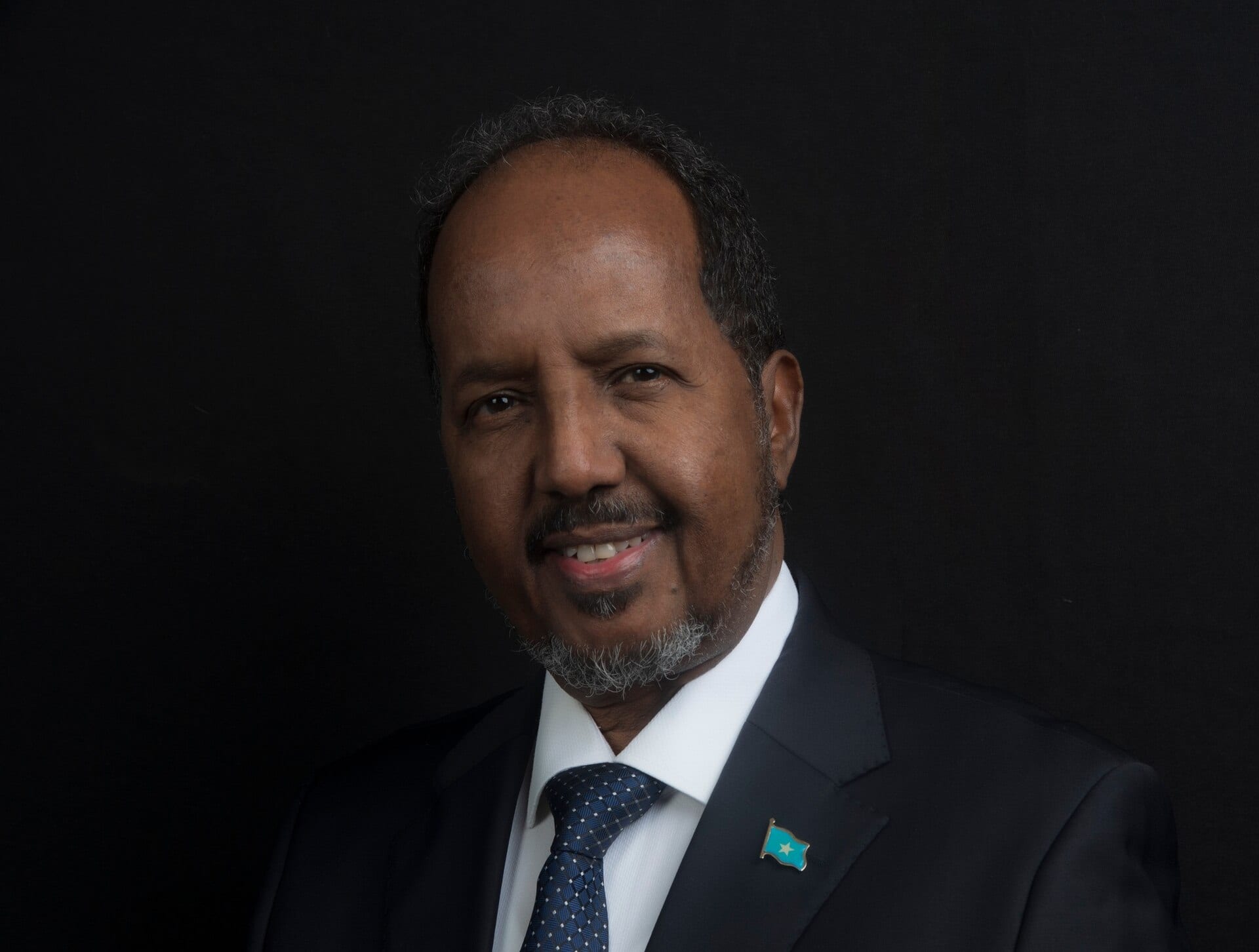 Official portrait of a man in a dark suit and blue tie with a small Somali flag pin on his lapel, against a black background.