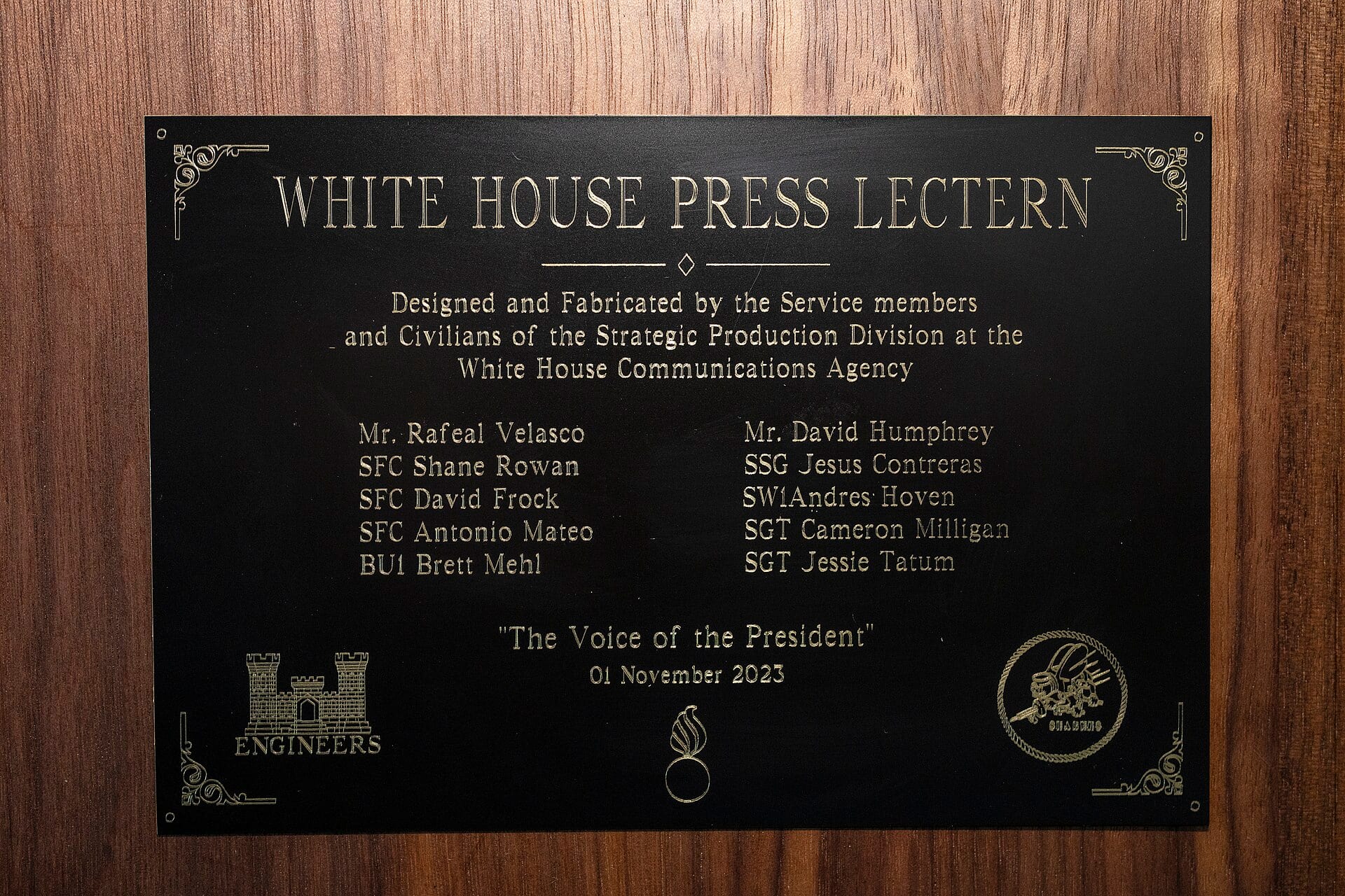 Placa negra con letras doradas que dice White House Press Lectern, montada sobre una pared de madera.