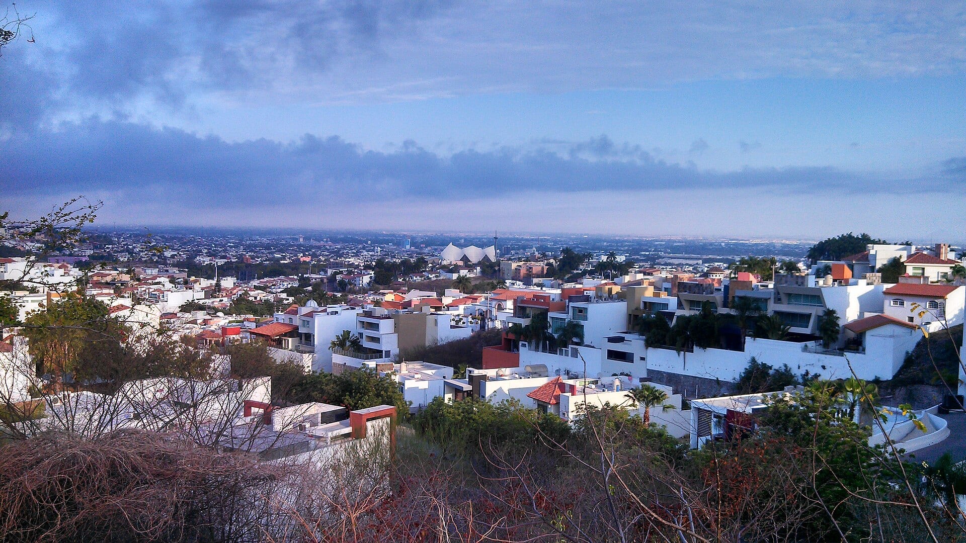 Vista de Culiacán desde un cerro, con casas blancas y de tejas rojas en primer plano y la ciudad extendida hasta el horizonte bajo un cielo nublado.