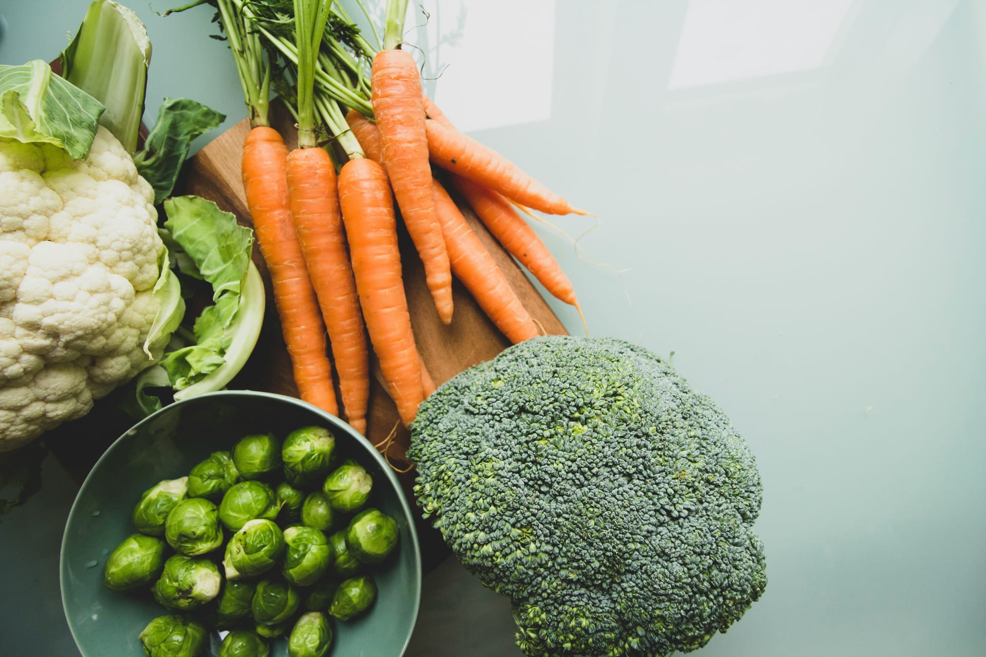 Zanahorias, brócoli, coliflor y coles de Bruselas sobre una tabla de madera y una superficie clara.