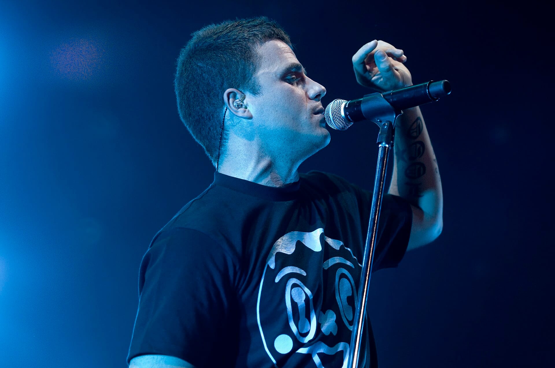 Dani Martín, de perfil y con el brazo levantado, canta ante un micrófono bajo luz azul durante un concierto de El Canto del Loco en 2008.