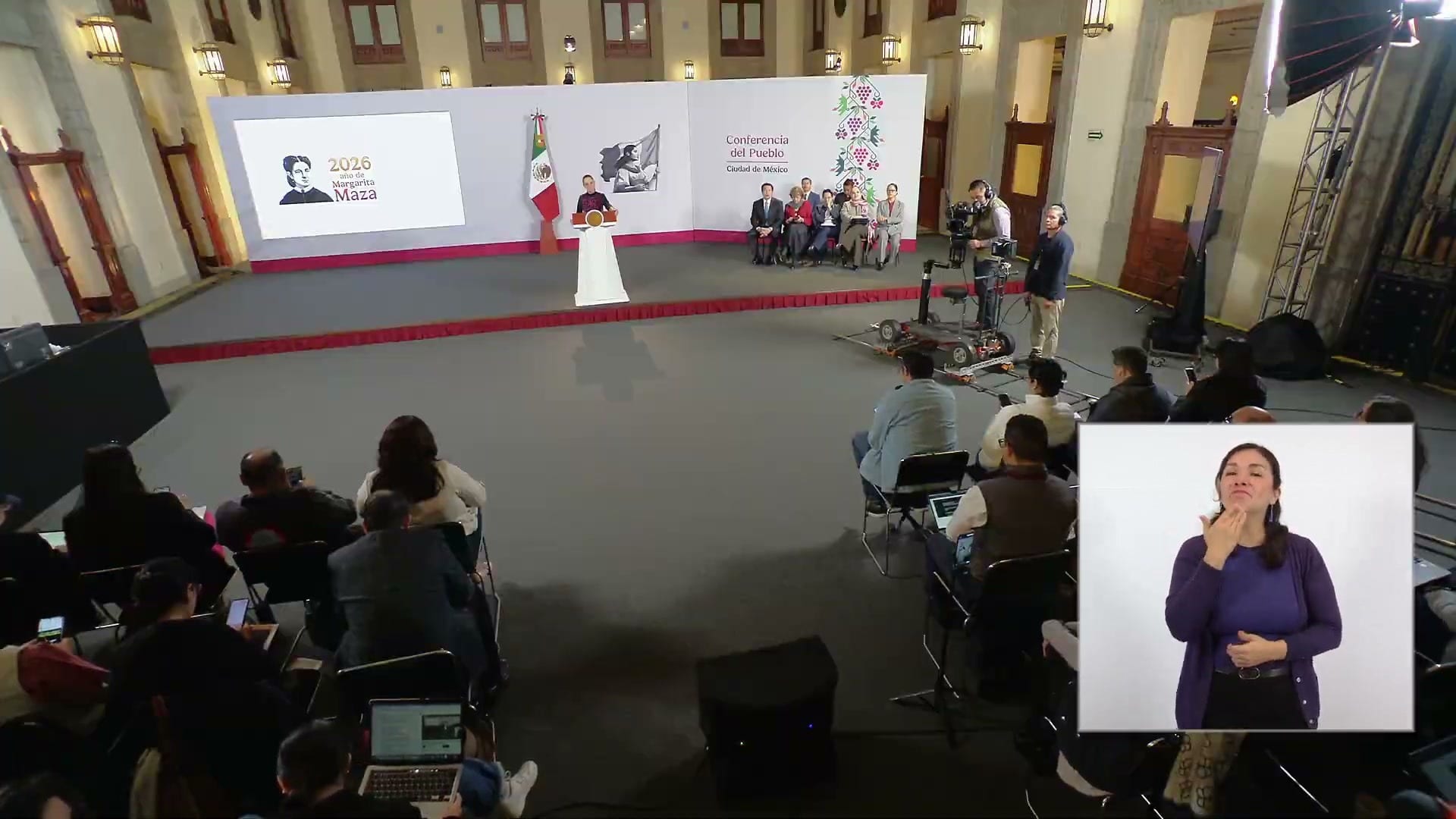 La presidenta habla desde el podio de la sala de conferencias, con reporteros sentados en primer plano y una intérprete de lengua de señas en un recuadro.