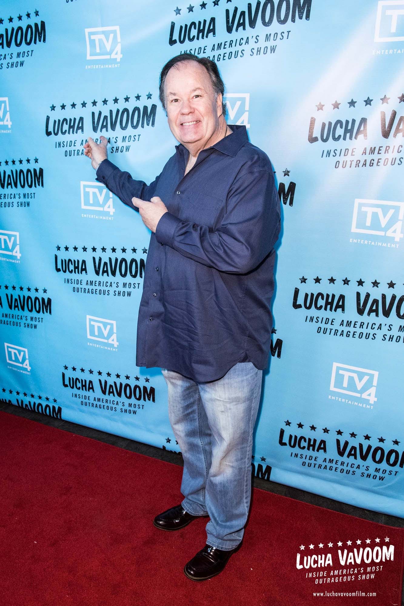 Hombre mayor de cabello canoso y camisa azul sonríe y señala un fondo de evento sobre una alfombra roja.