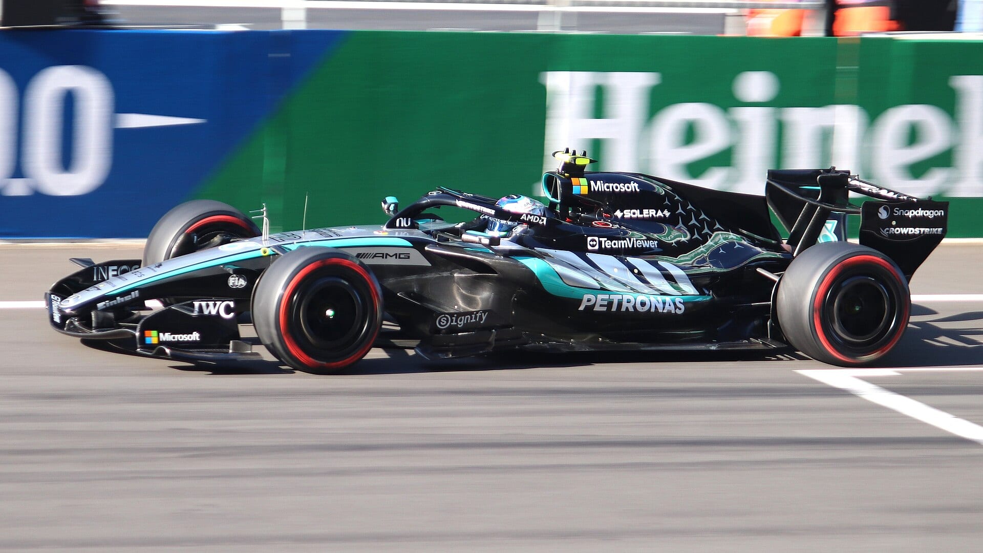 A black and silver Mercedes F1 car with red-striped tires speeds past a blue and green advertising wall on a racetrack.