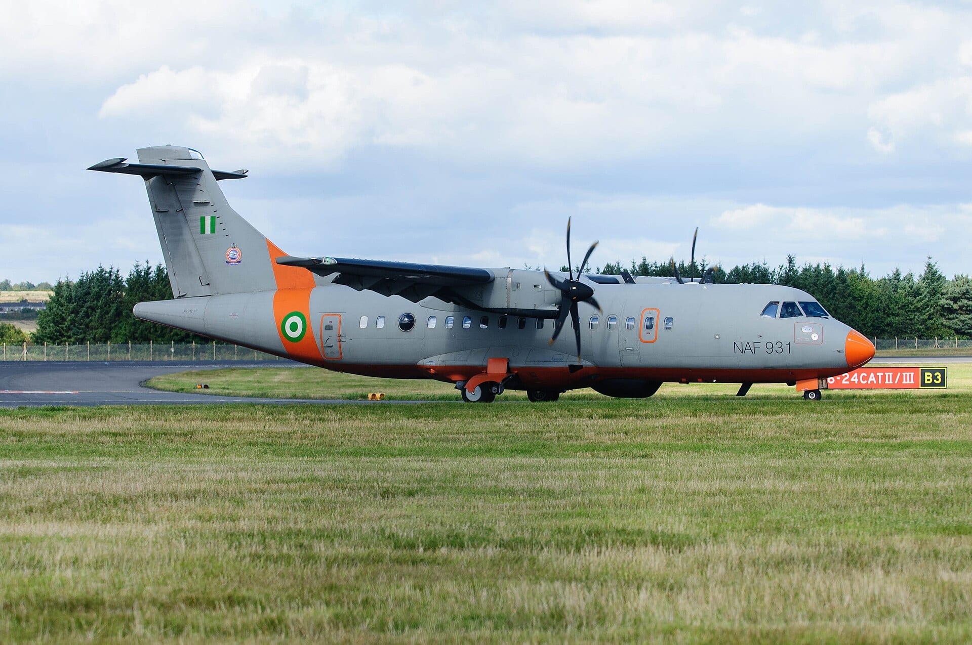 A gray turboprop plane with an orange nose and the marking NAF 931 taxis on a runway under a cloudy sky.