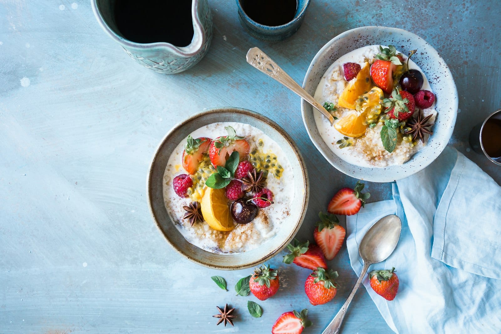 Dos tazones de avena con fresas, frambuesas, naranja y una cereza, junto a una cuchara y una servilleta azul sobre una mesa.