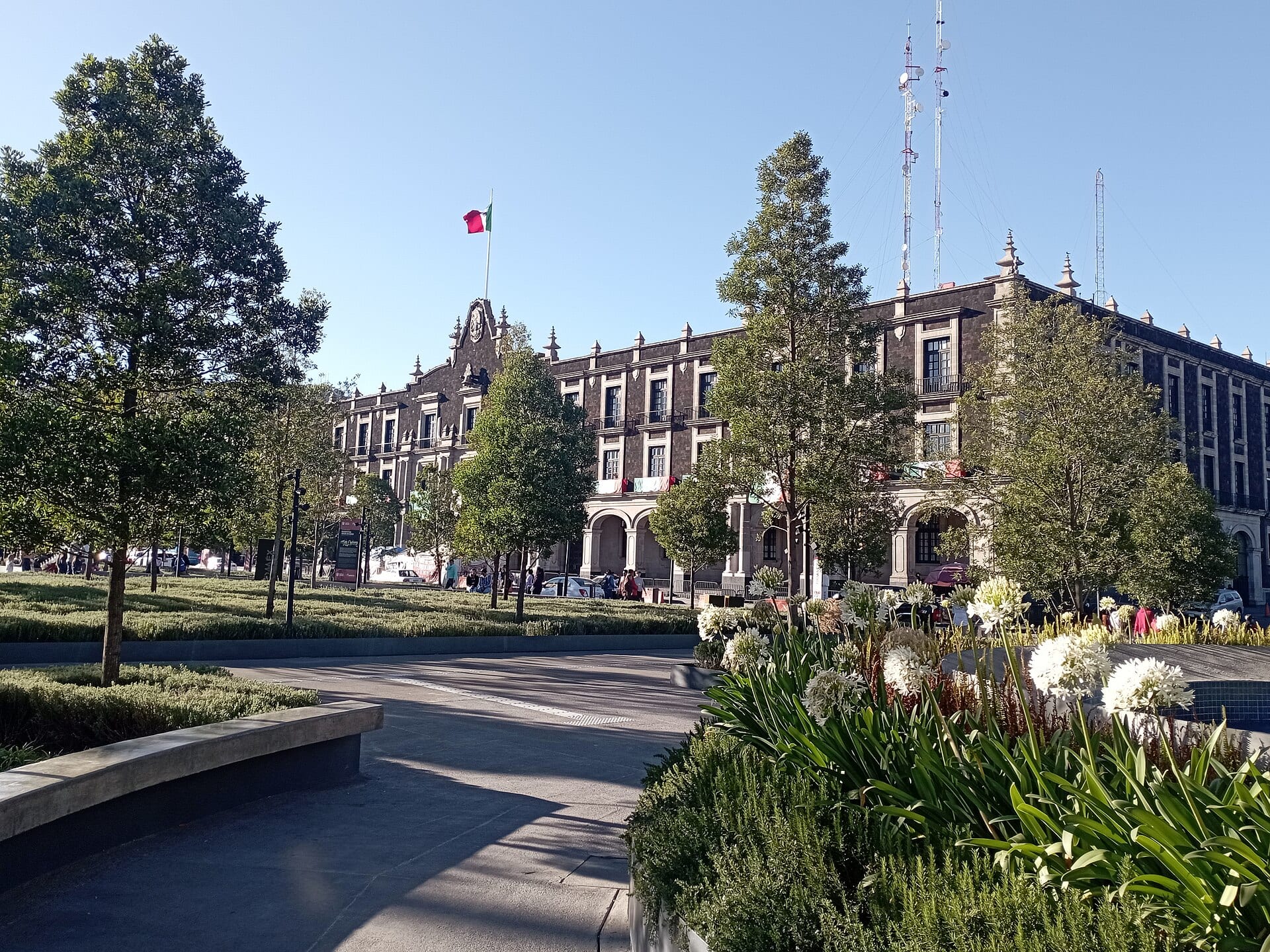 Fachada de piedra oscura del Palacio de Gobierno de Toluca con una bandera de México, vista desde una plaza con árboles y flores blancas.