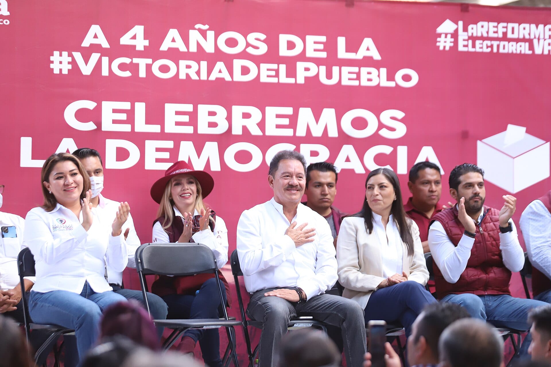 Un hombre de camisa blanca sentado con la mano en el pecho, entre otras personas, frente a una lona roja con el lema Celebremos la democracia.