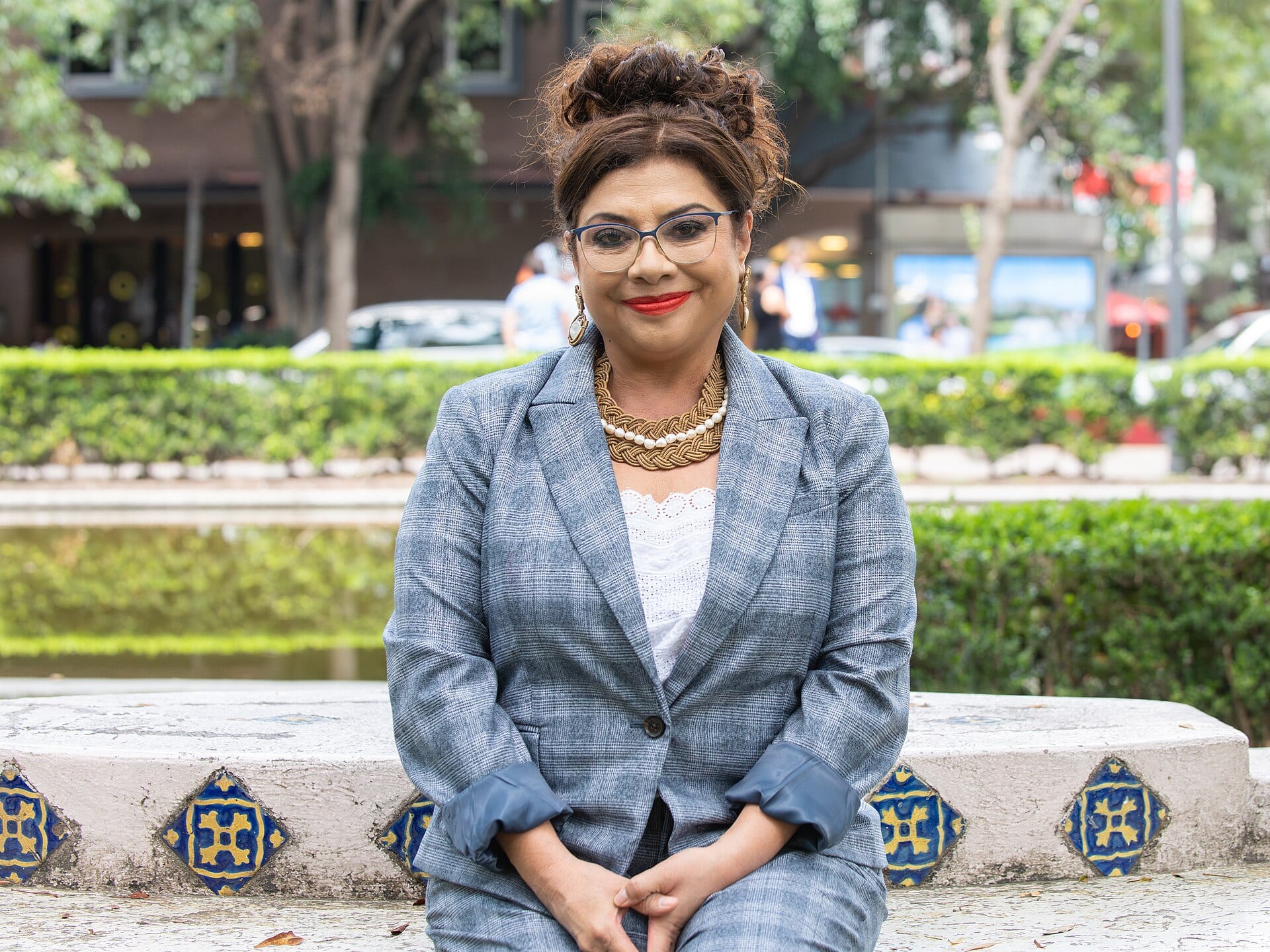 Una mujer con saco gris y lentes sonríe sentada en una banca con azulejos azules, frente a un seto y un estanque.