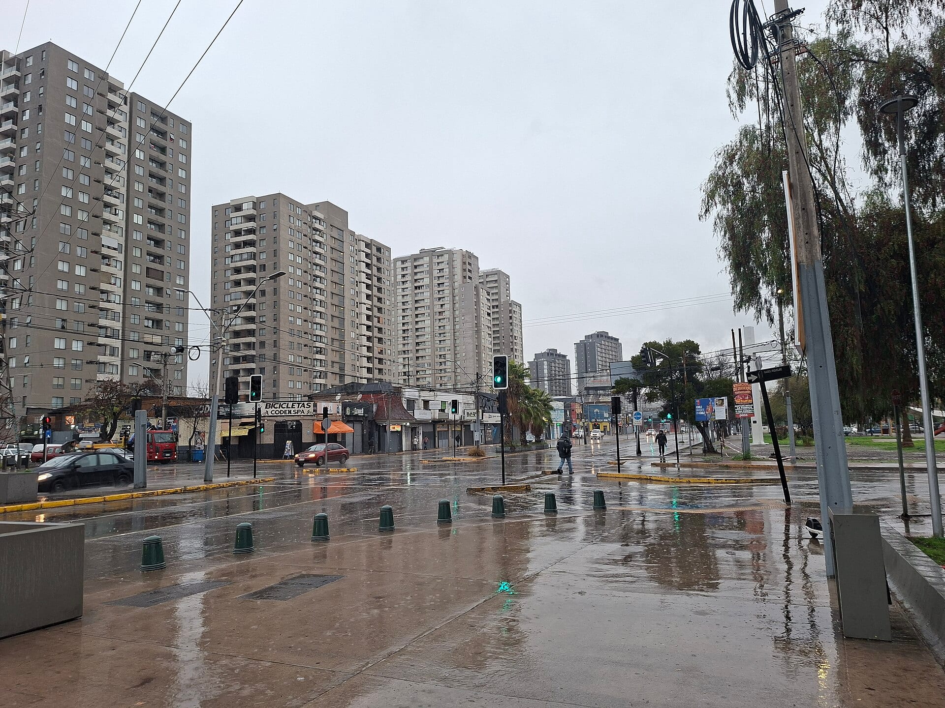 Una avenida mojada de Santiago bajo la lluvia, con torres de departamentos al fondo y peatones cruzando entre semáforos.