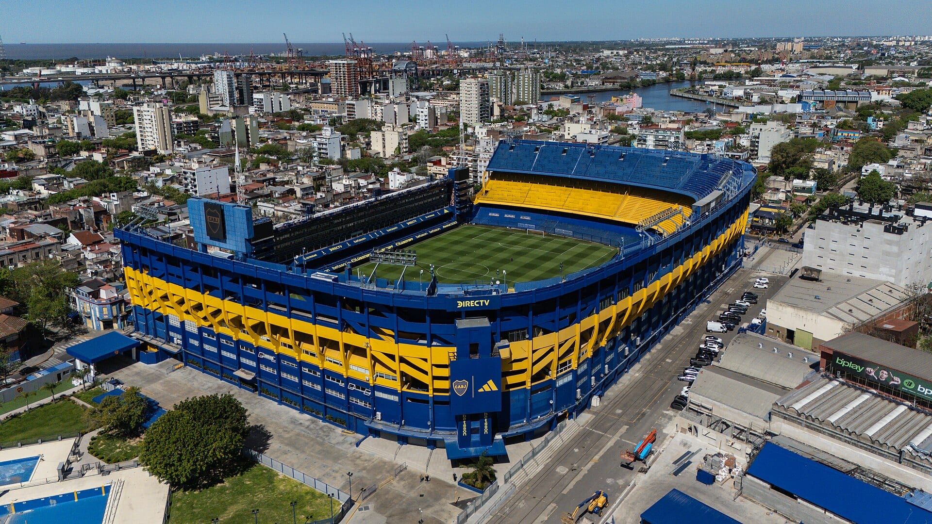 Vista aérea de La Bombonera, con sus tribunas azules y amarillas alrededor de la cancha, y las grúas del puerto al fondo.