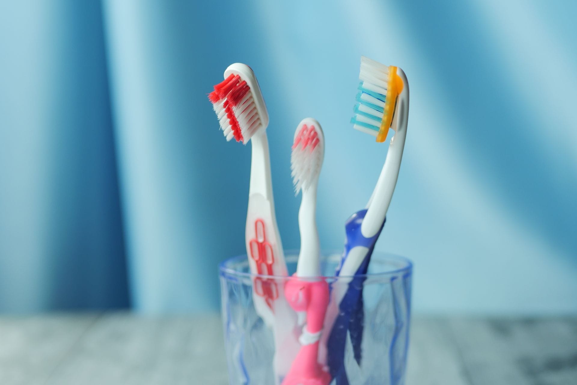 Tres cepillos de dientes, uno rojo, uno rosa y uno azul con amarillo, dentro de un vaso transparente frente a una tela azul.