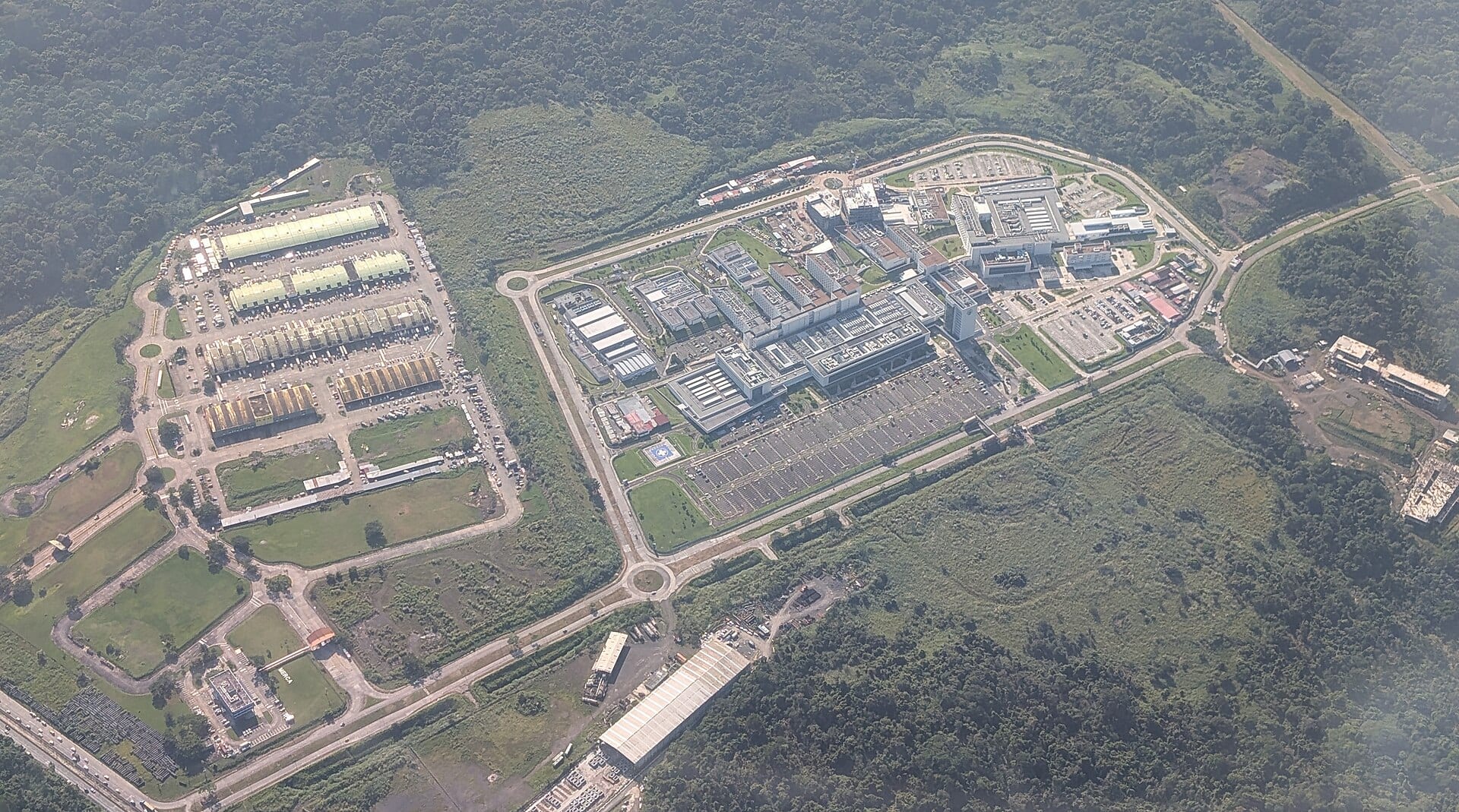 Aerial view of a hospital complex of white buildings with parking lots and a helipad, surrounded by forest.