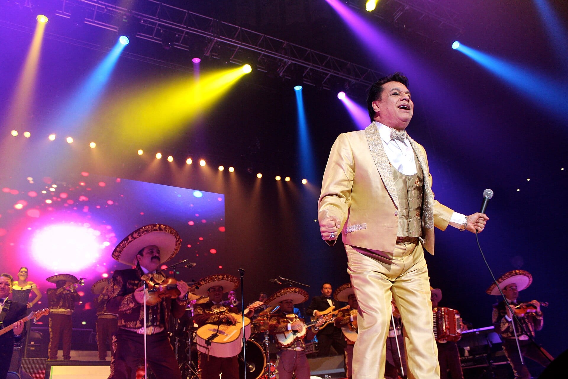 Juan Gabriel, de traje dorado y micrófono en mano, canta en un escenario con mariachi y luces de colores.