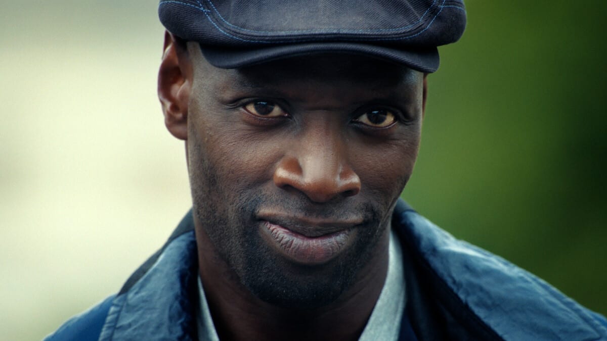 Primer plano de Omar Sy, con gorra oscura y chamarra azul, que mira a la cámara con una leve sonrisa.