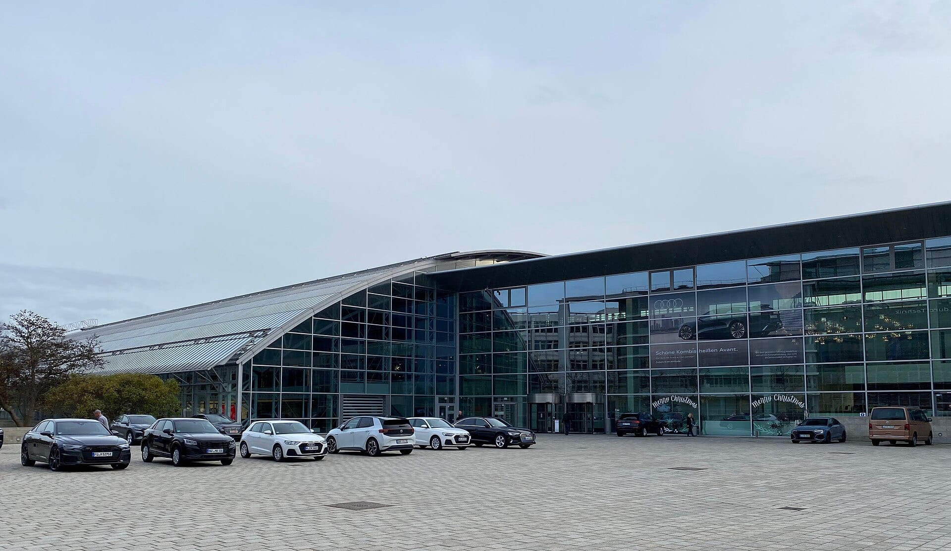 Fachada de vidrio con techo curvo del Audi Forum en Ingolstadt, con varios autos de la marca estacionados frente a la entrada.