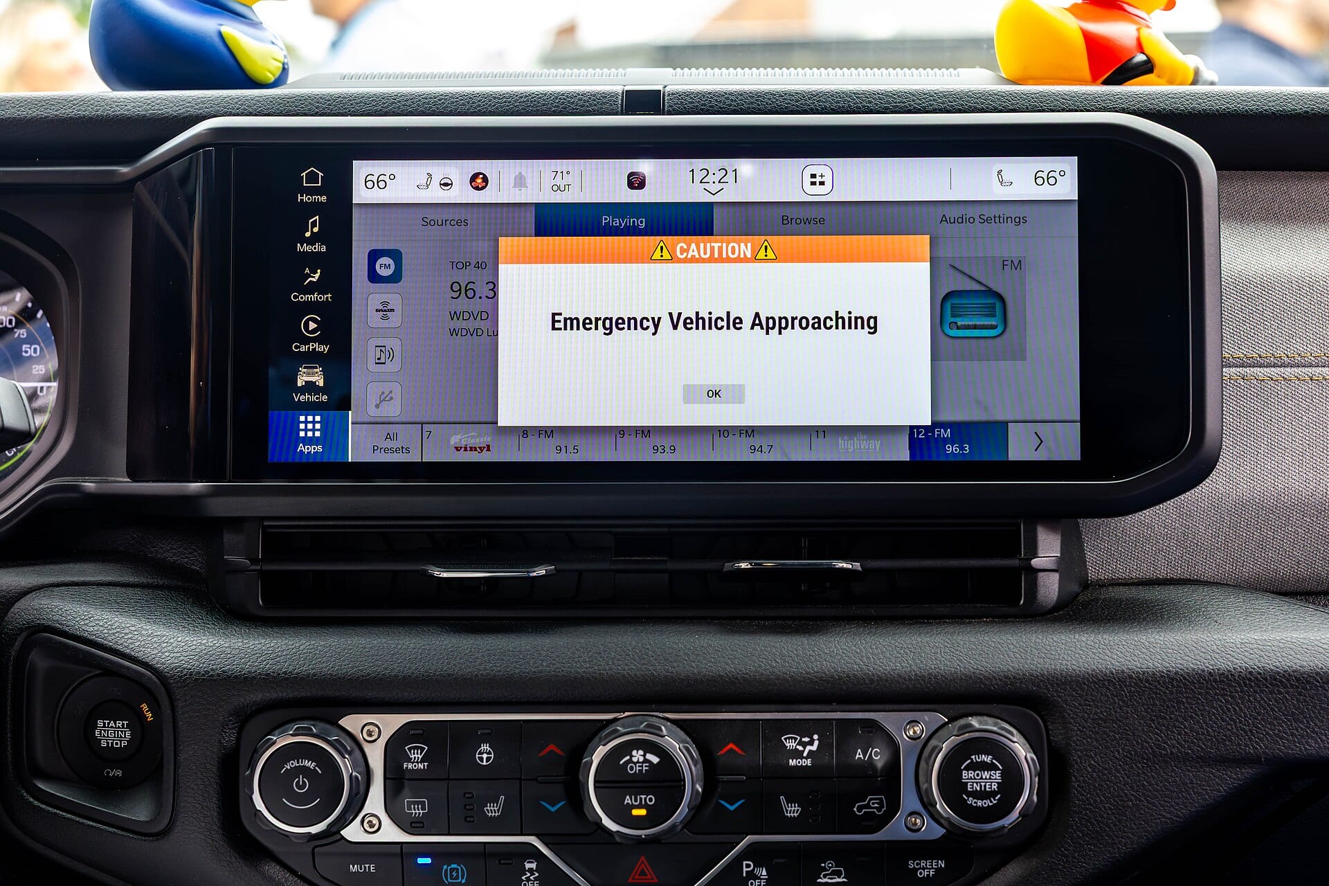 Pantalla de infoentretenimiento de un auto con un aviso de precaución que dice Emergency Vehicle Approaching, sobre el tablero y los controles de clima.