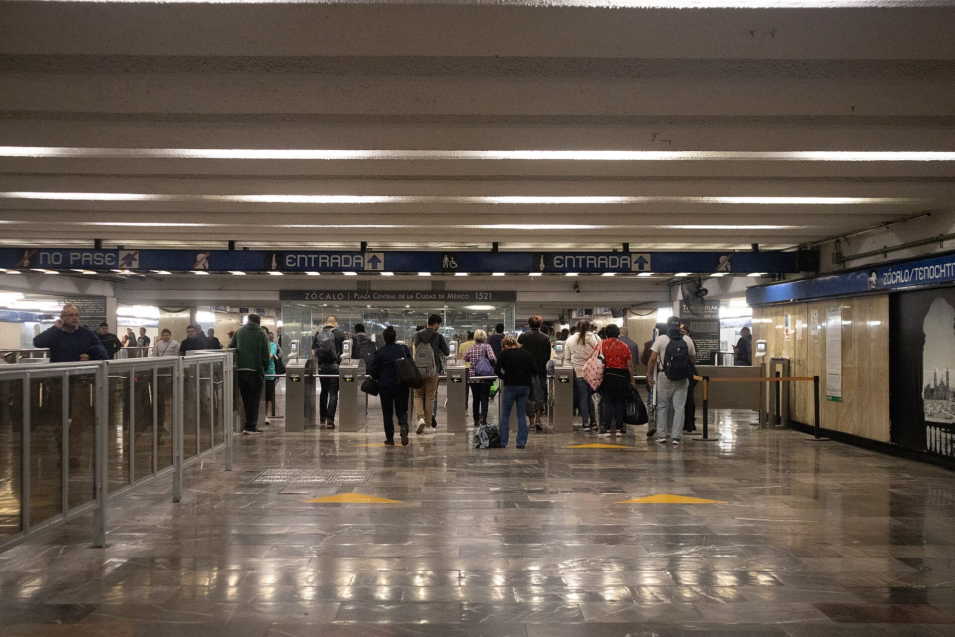 Usuarios con mochilas y bolsas cruzan los torniquetes de un acceso del Metro, bajo un letrero azul de entrada y el rótulo de Zócalo.