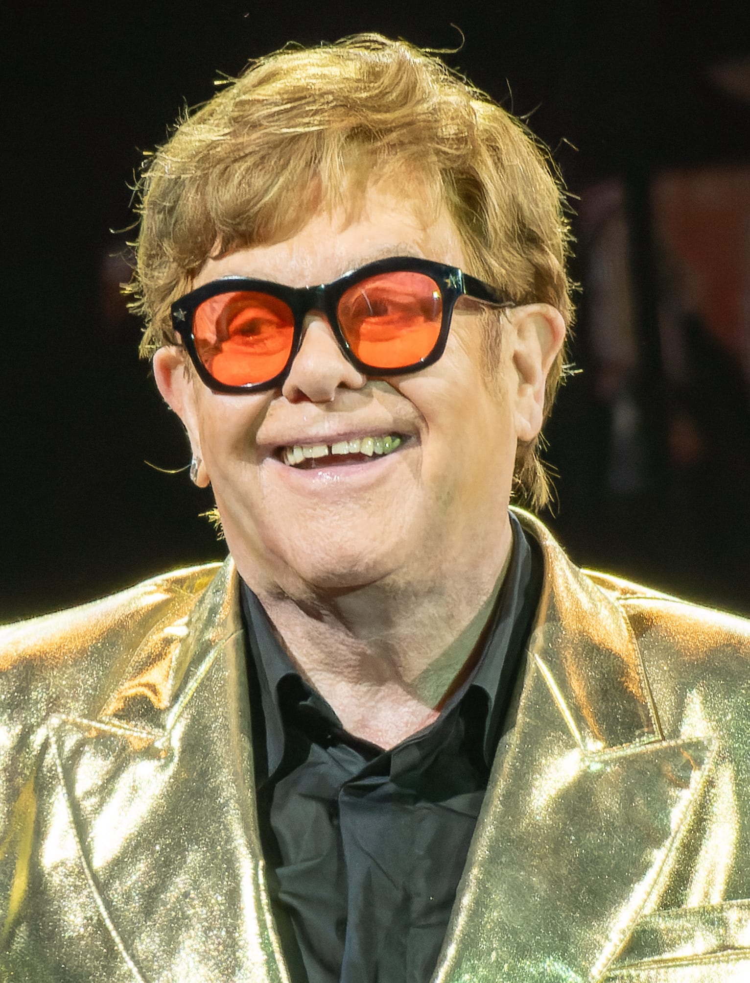 Elton John sonríe con lentes de armazón negro y cristales naranjas, y un saco dorado brillante sobre camisa negra, en un escenario.