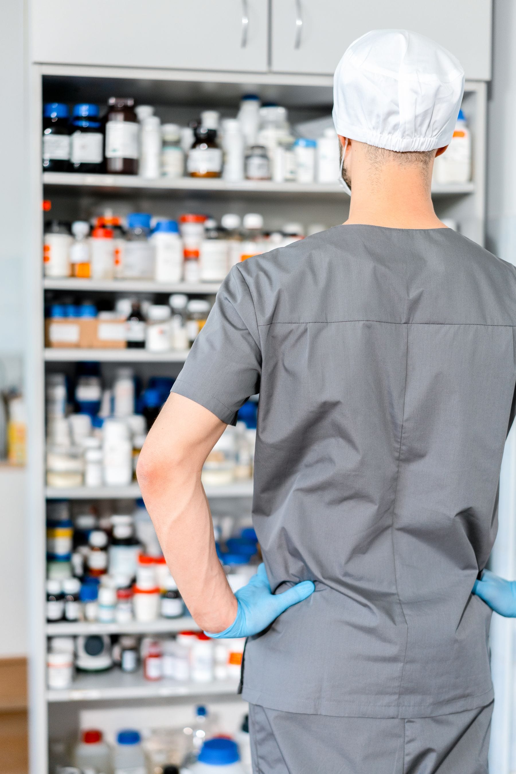 Persona con gorro blanco, bata gris y guantes azules, vista de espaldas, frente a un estante lleno de frascos y botes de medicamentos.