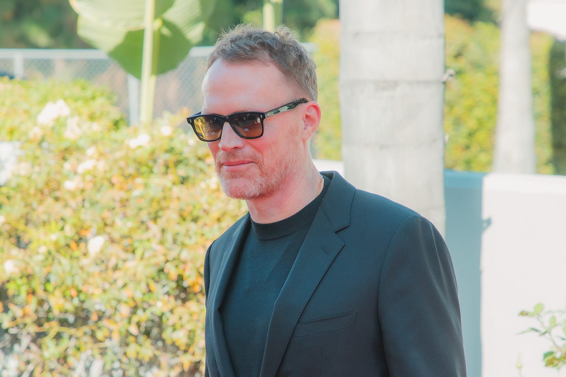 Paul Bettany, con lentes oscuros y saco negro, posa de perfil frente a unos arbustos con flores blancas durante una alfombra roja.