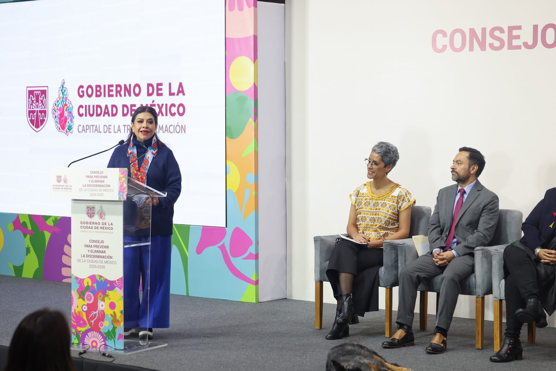 Clara Brugada habla en un podio con el letrero del Consejo para Prevenir y Eliminar la Discriminación; a su derecha, tres personas escuchan sentadas.