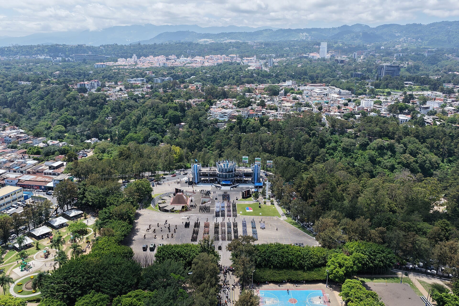 Aerial view of the Campo Marte complex in Guatemala City, with a parade ground surrounded by trees and the city in the background.