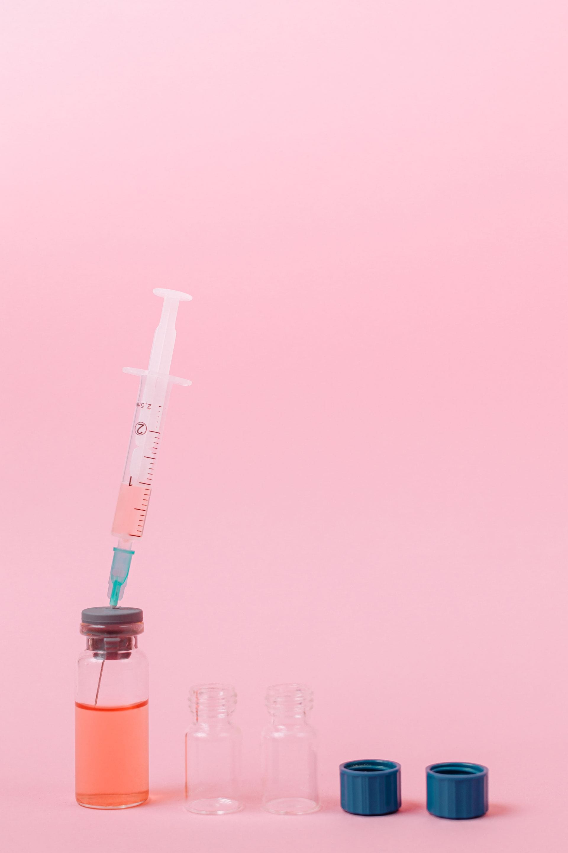 A syringe with pink liquid stuck into a glass vial, next to two empty vials and two blue caps on a pink background.