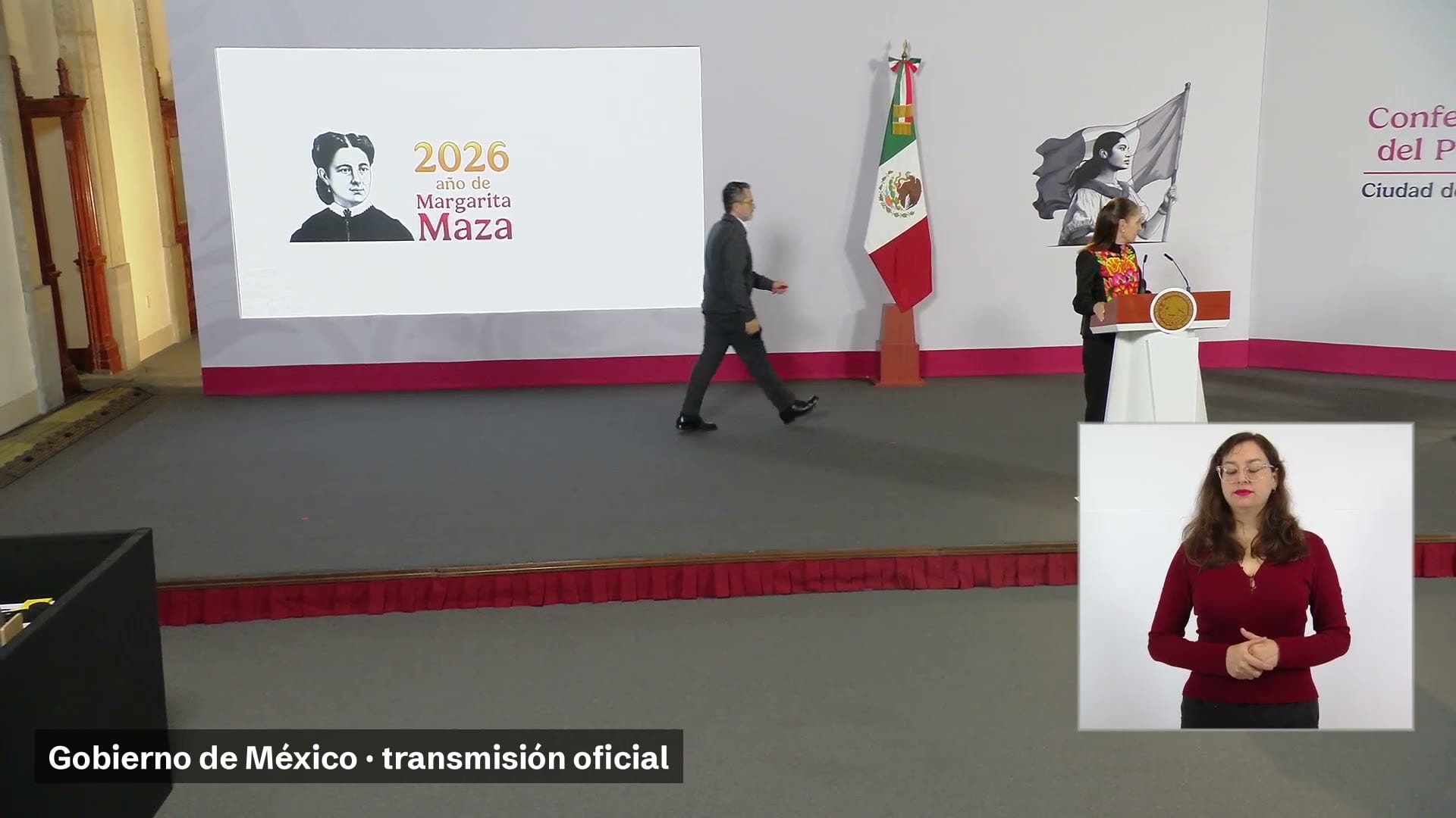 Una mujer de blusa bordada de colores en el podio de la sala de conferencias, un hombre que camina frente a una pantalla y una intérprete de lengua de señas.