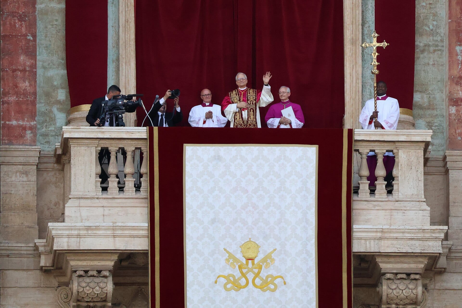 El papa León XIV saluda con la mano desde el balcón central de la Basílica de San Pedro, flanqueado por ayudantes, con un camarógrafo a su izquierda.