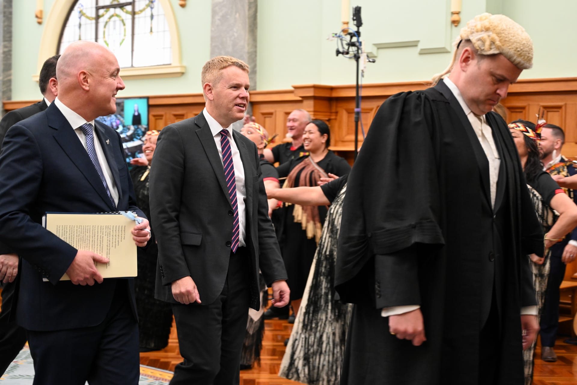 Christopher Luxon, a la izquierda, y Chris Hipkins, ambos de traje, caminan por un salón del Parlamento junto a un hombre con toga y peluca y un grupo de danzantes maoríes.