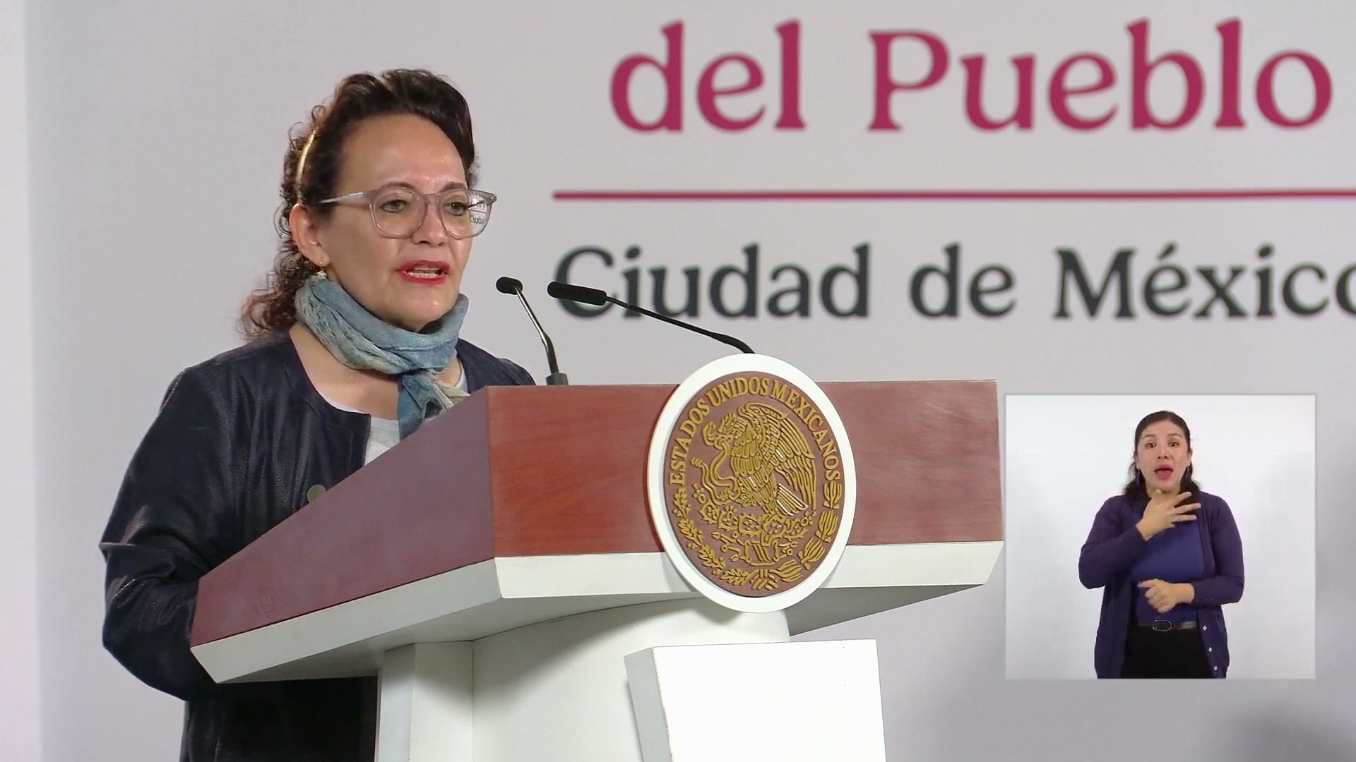 Una mujer con lentes habla desde el podio de la conferencia matutina; a la derecha, una intérprete de lengua de señas.