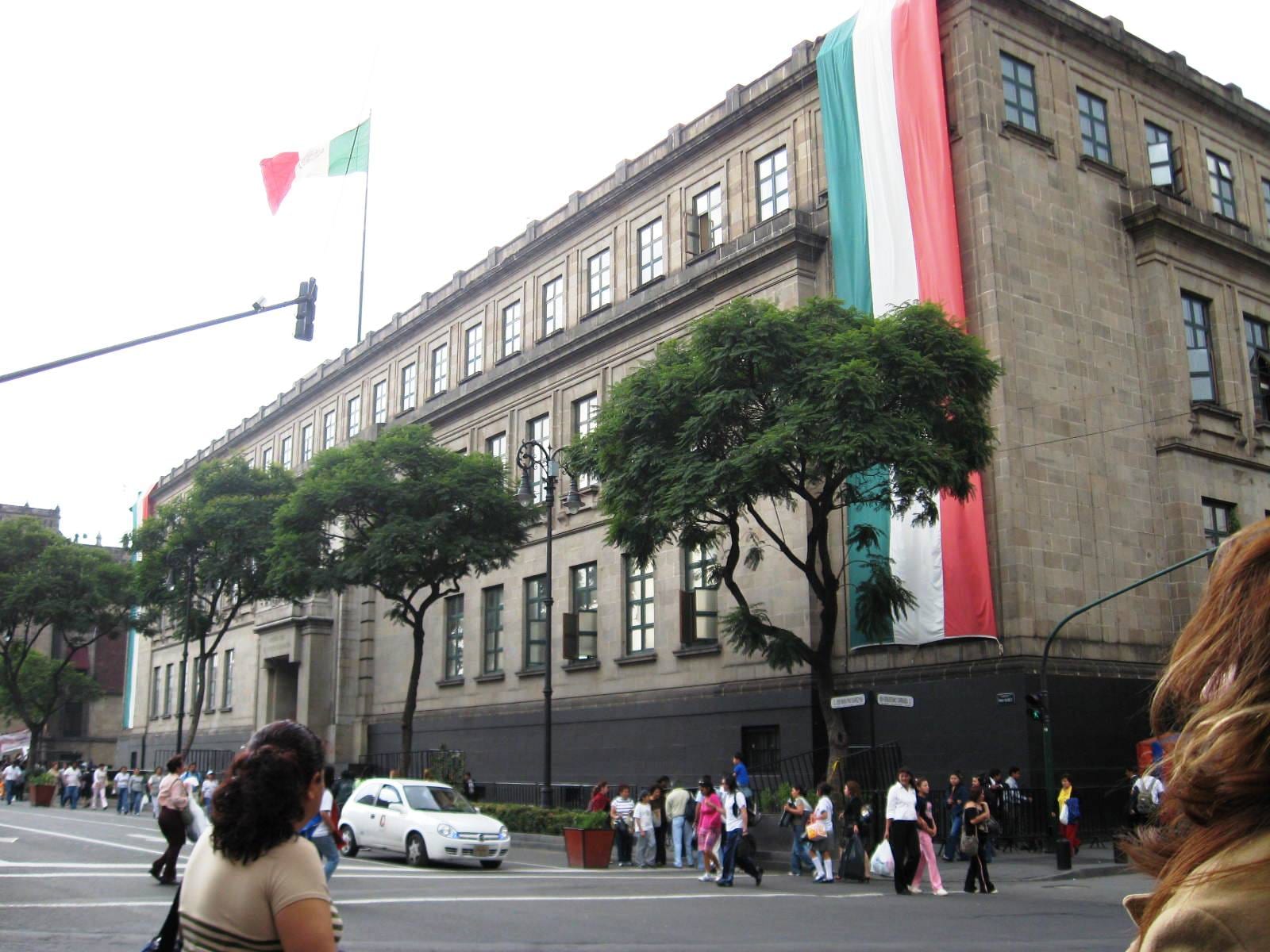 Edificio de piedra gris de la Suprema Corte de Justicia en la Ciudad de México, con una bandera tricolor gigante colgada de la esquina y gente cruzando la calle.