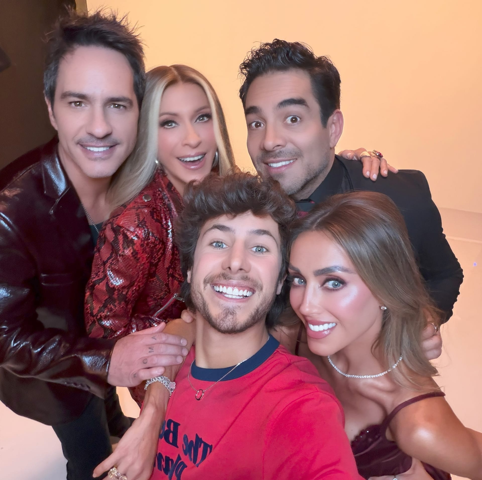 Cinco personas sonríen apretadas en un selfie grupal sobre un fondo anaranjado; en primer plano, un joven con camiseta roja.