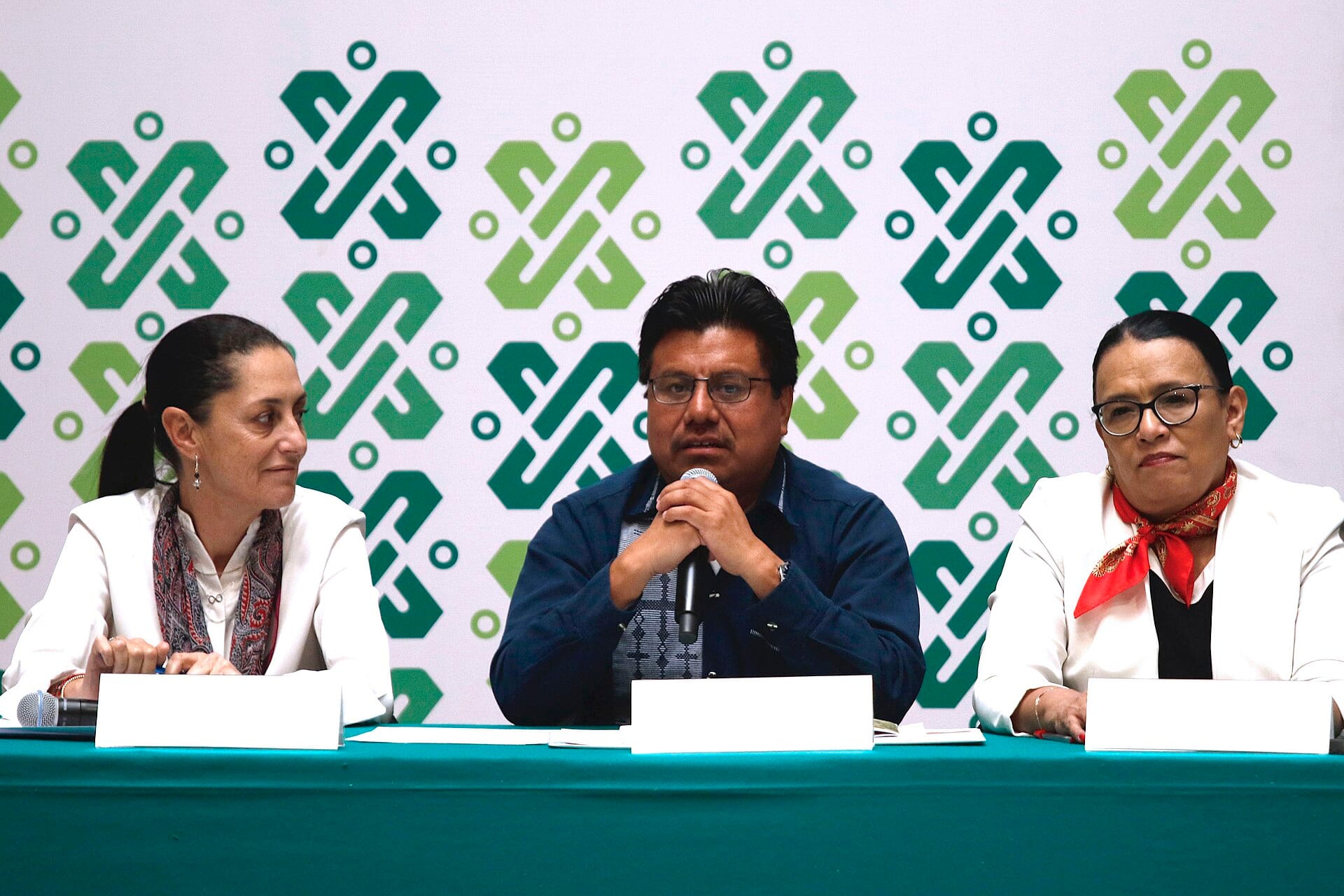 Tres personas sentadas a una mesa con mantel verde, frente a un fondo de motivos geométricos verdes; la del centro habla por un micrófono.