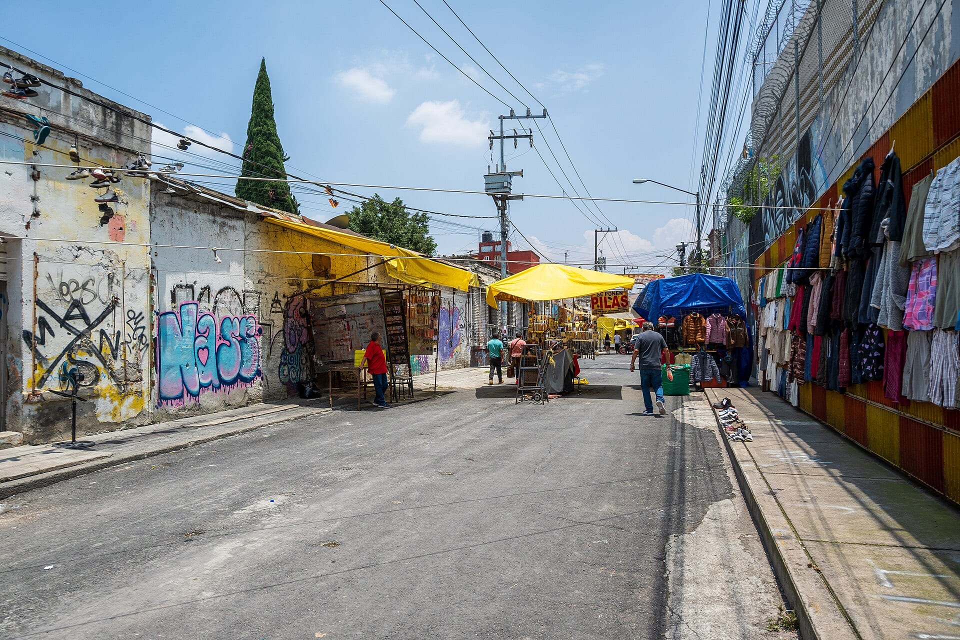 Calle de Tepito con puestos bajo toldos amarillos y azules, y ropa colgada de una pared a la derecha.