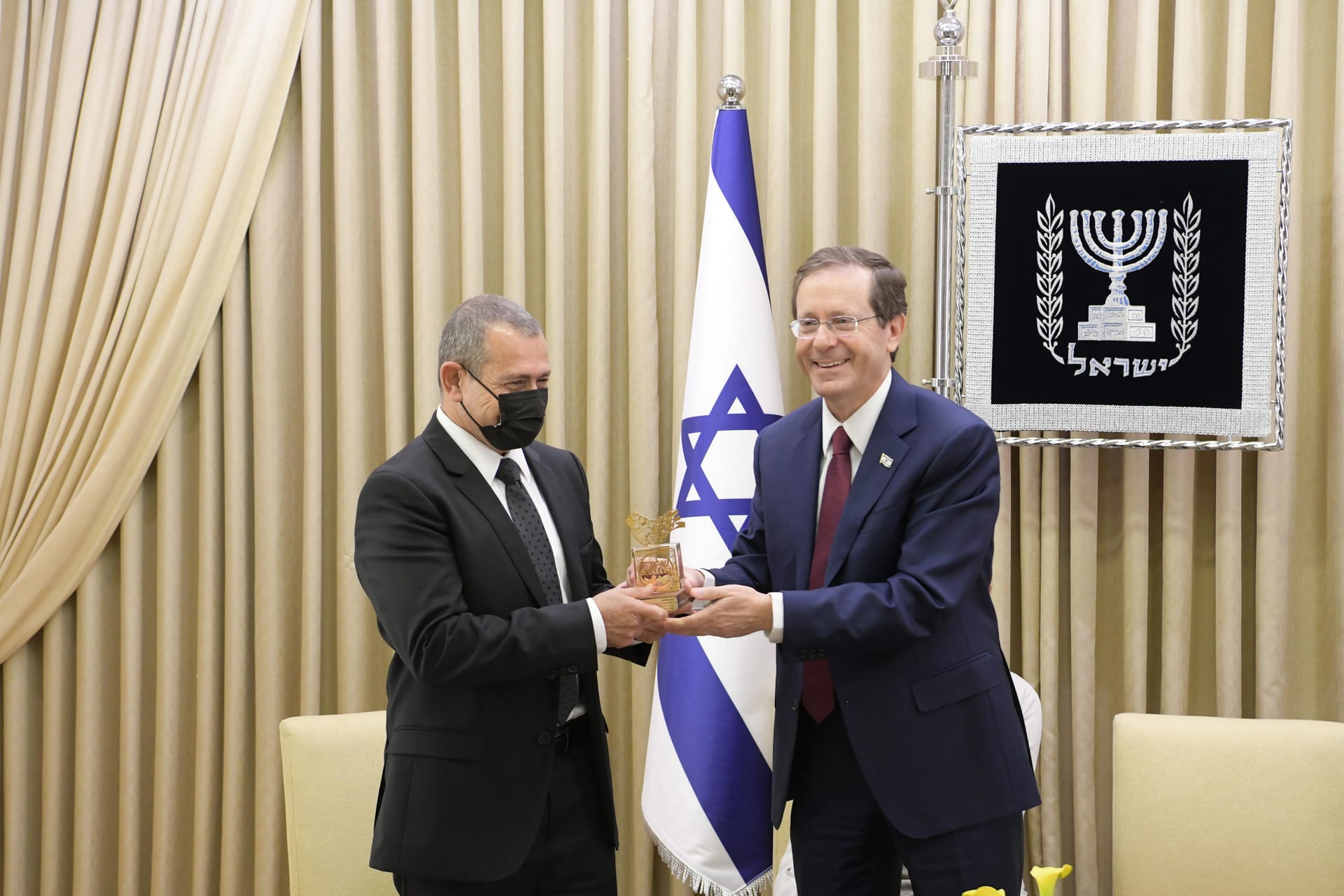 Dos hombres de traje, uno de ellos con cubrebocas negro, sostienen juntos un trofeo de cristal frente a una bandera de Israel y cortinas doradas.