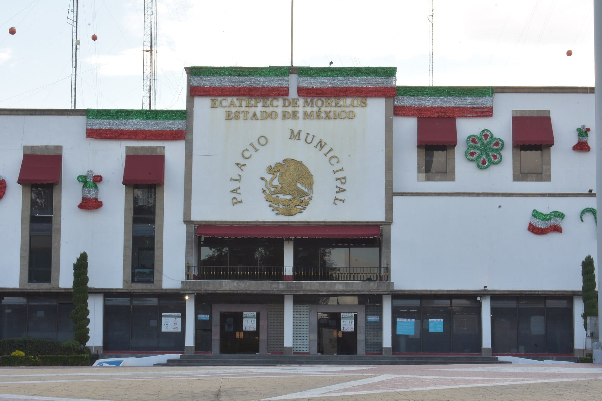 Fachada blanca del Palacio Municipal de Ecatepec con el escudo nacional dorado, adornos tricolores y una explanada de adoquín al frente.