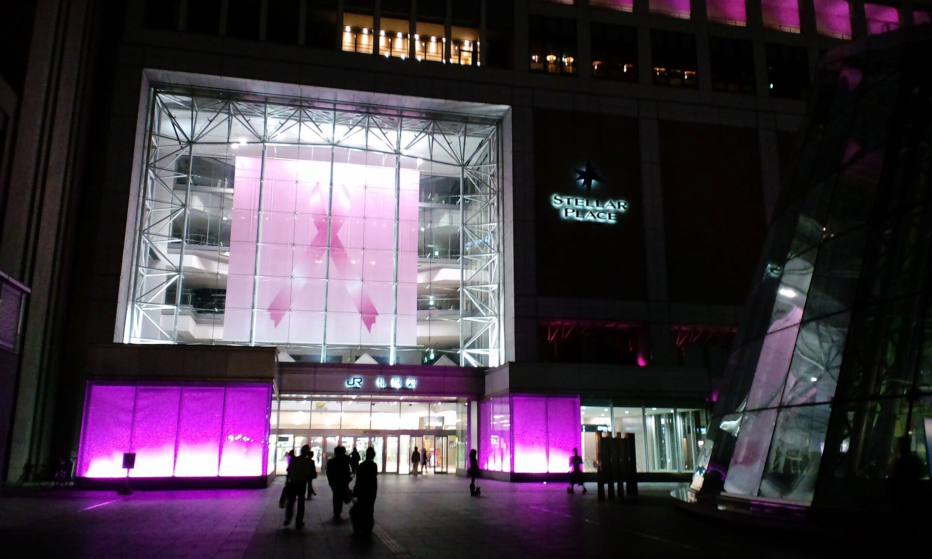 Fachada iluminada de noche con un gran listón rosa de la lucha contra el cáncer de mama en una ventana de cristal.