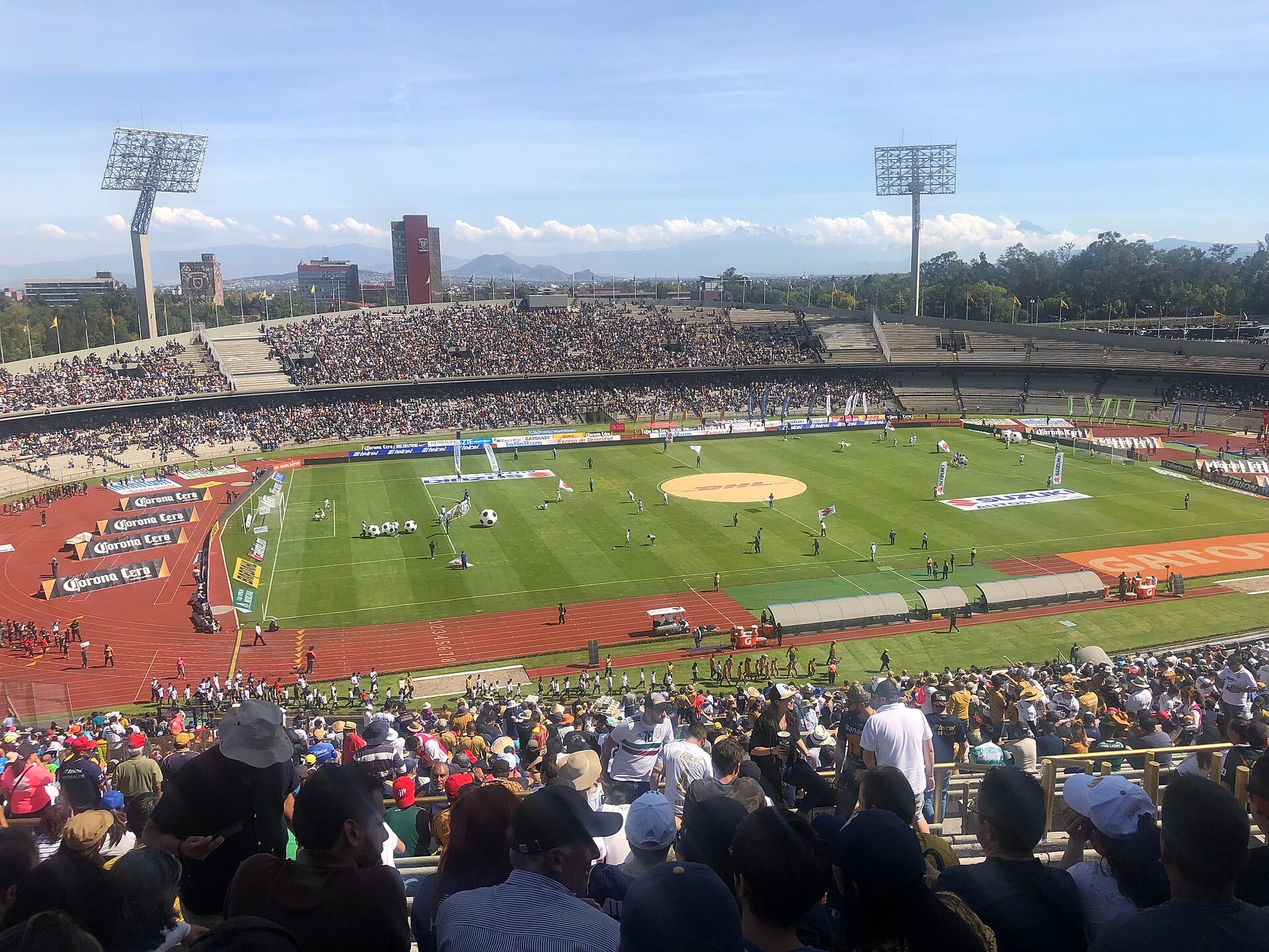 Vista del Estadio Olímpico Universitario desde la tribuna, con el campo, la pista roja y las gradas llenas bajo un cielo despejado.