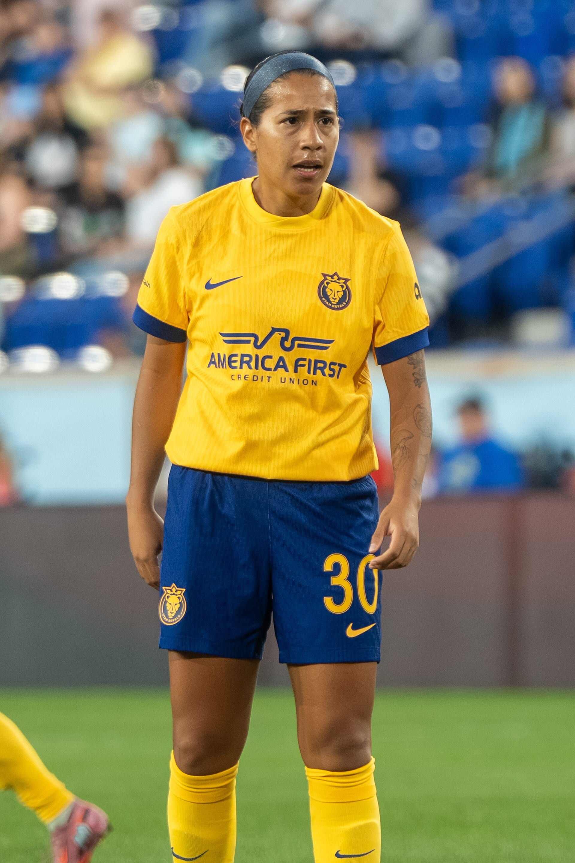 Una futbolista con playera amarilla y short azul con el número 30 está de pie sobre el pasto de un estadio.