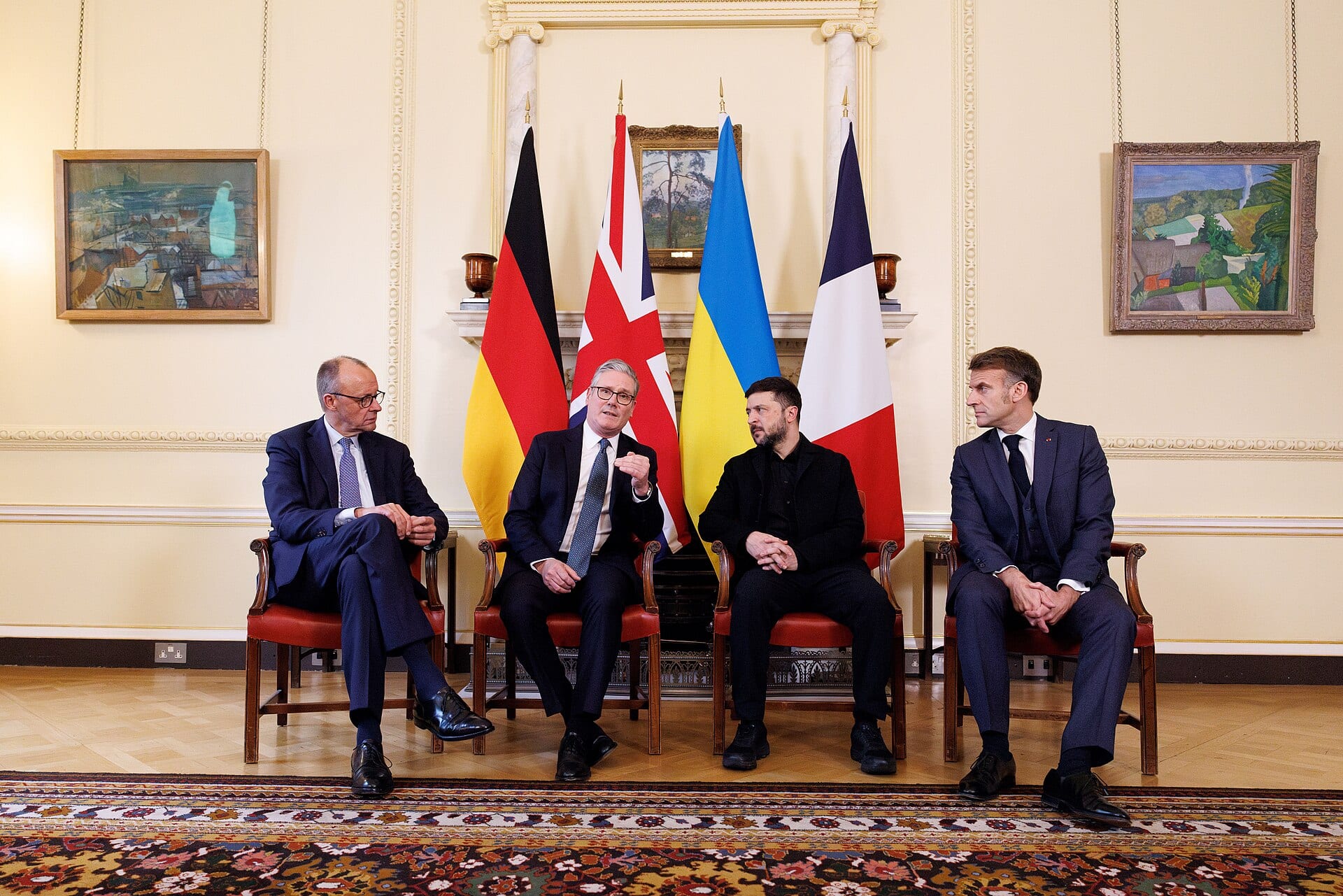Four men sit on red chairs in front of the flags of Germany, the United Kingdom, Ukraine and France in a light-walled room.