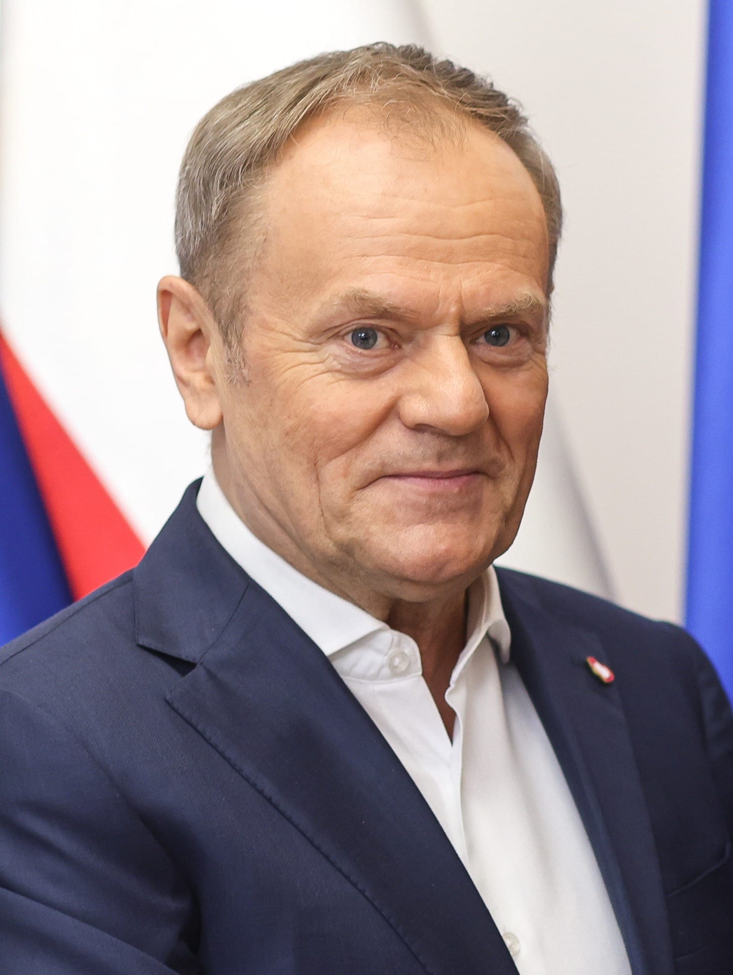 Portrait of Donald Tusk in a navy suit and white shirt, standing in front of flags.