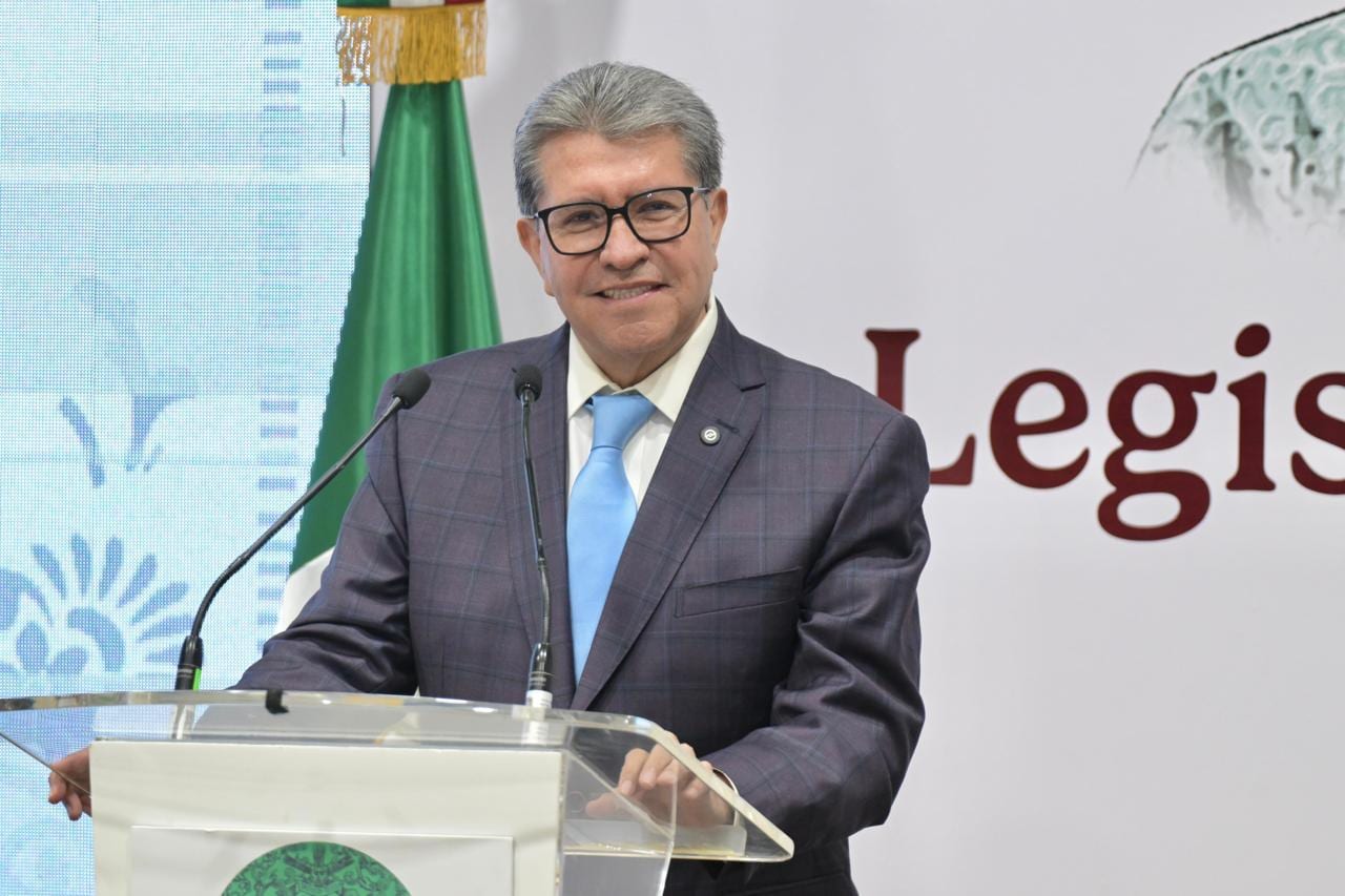 Ricardo Monreal, de saco gris, corbata azul y lentes, habla desde un podio de acrílico con la bandera de México al fondo.